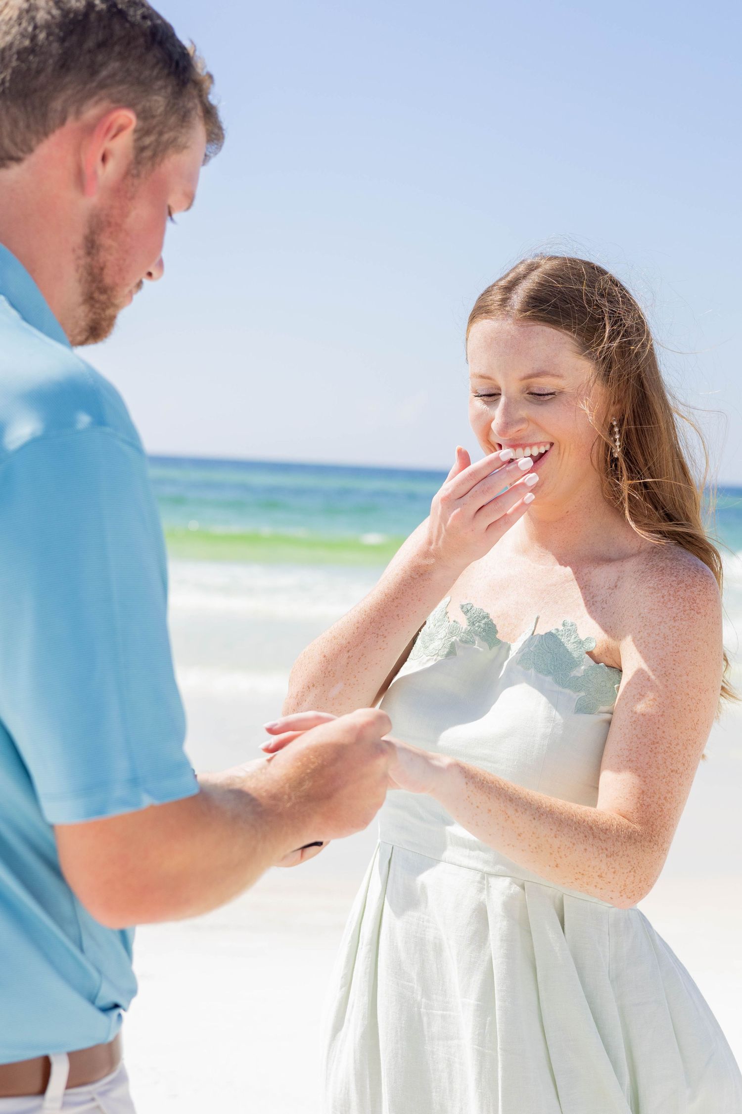 Man sliding engagement ring onto womans finger as she covers her mouth in surprise on the beach.