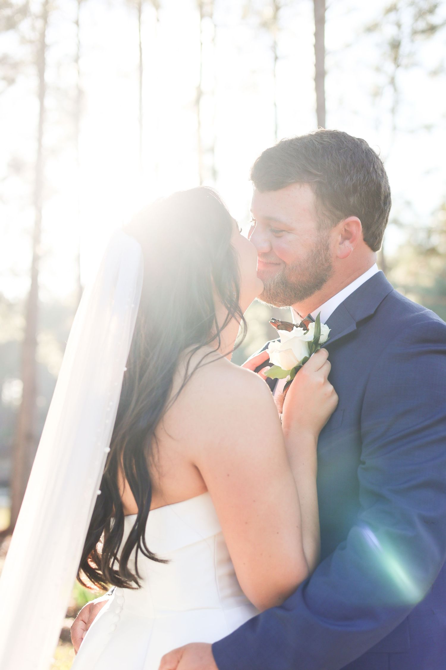 Newlywed couple shares romantic kiss in sunlit outdoor setting with white curtains and natural backdrop.