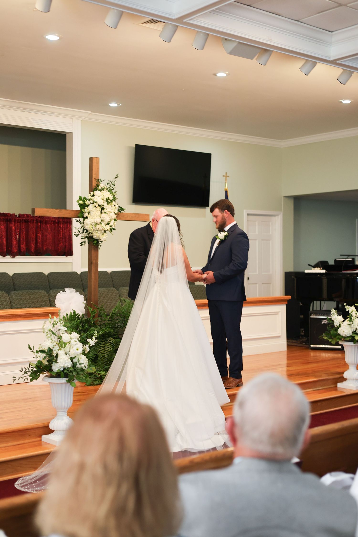 Wedding ceremony in a church sanctuary with white floral arrangements and guests seated in wooden pews.