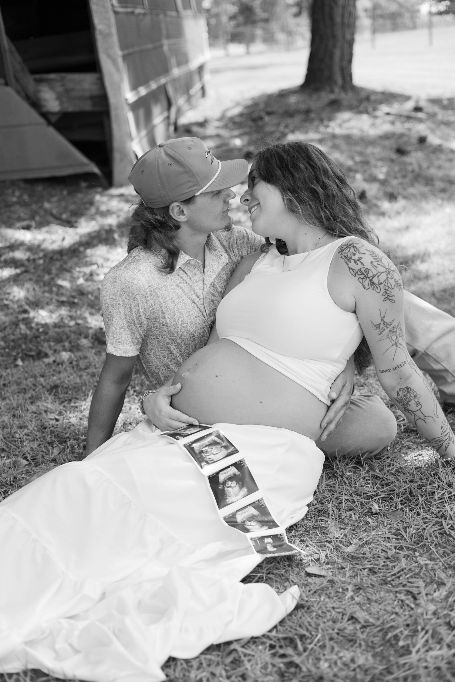 Black and white photo of a pregnant couple sitting on grass, sharing a tender moment with ultrasound photos displayed nearby.