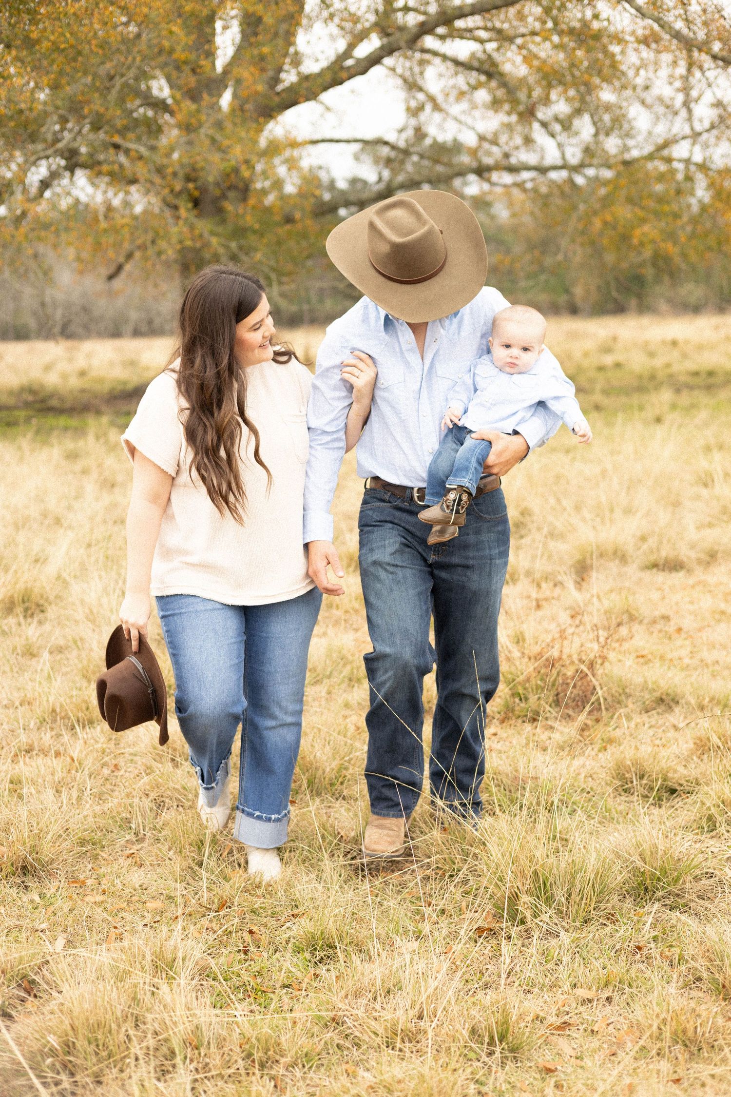 A young family walks through an autumn field, father in cowboy hat holding baby, mother beside him.