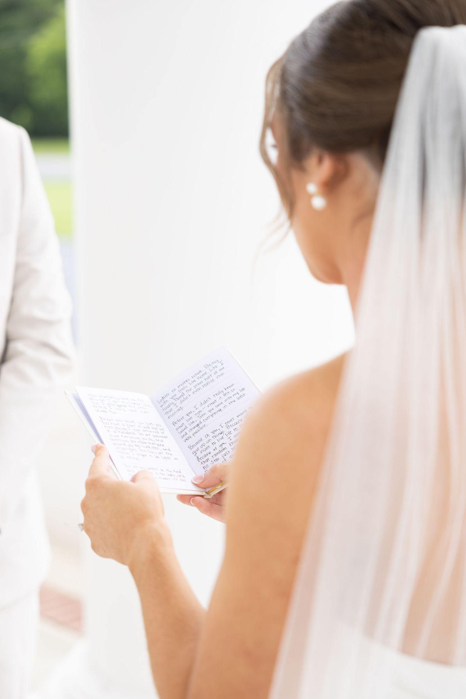 Bride in white veil reading handwritten wedding vows during outdoor ceremony.