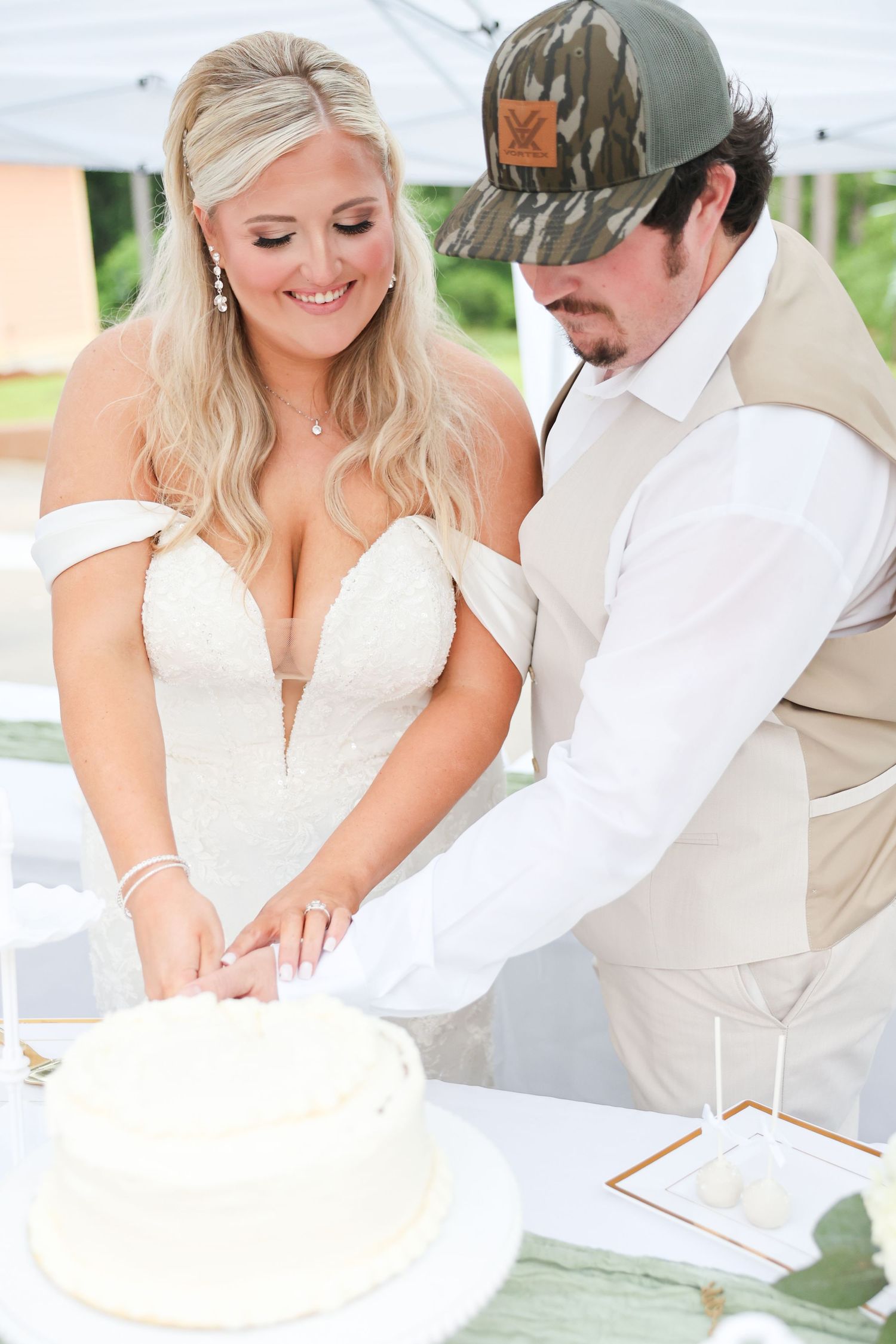 A smiling bride and groom in a camo cap cut their elegant white wedding cake together under a tent.