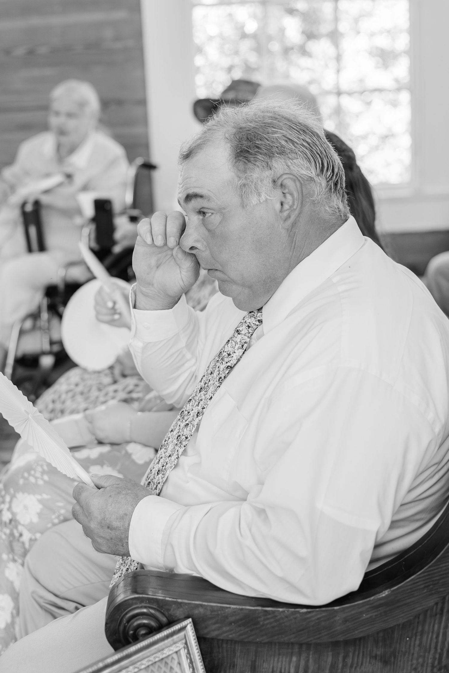 Emotional older man in white shirt and floral tie wiping tears during wedding ceremony in black and white.