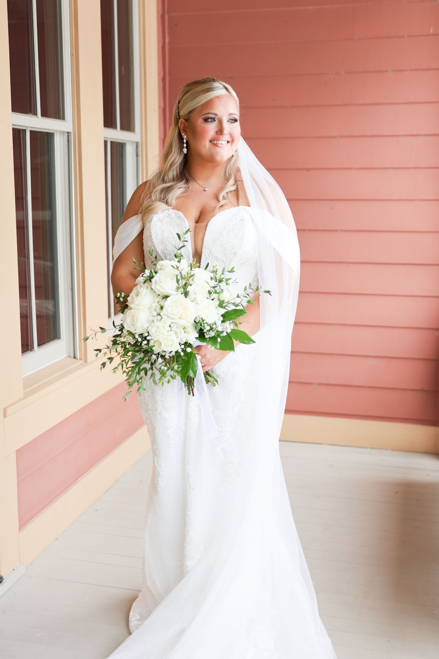 Beautiful bride in white lace mermaid gown holding white floral bouquet with veil outside pink building.