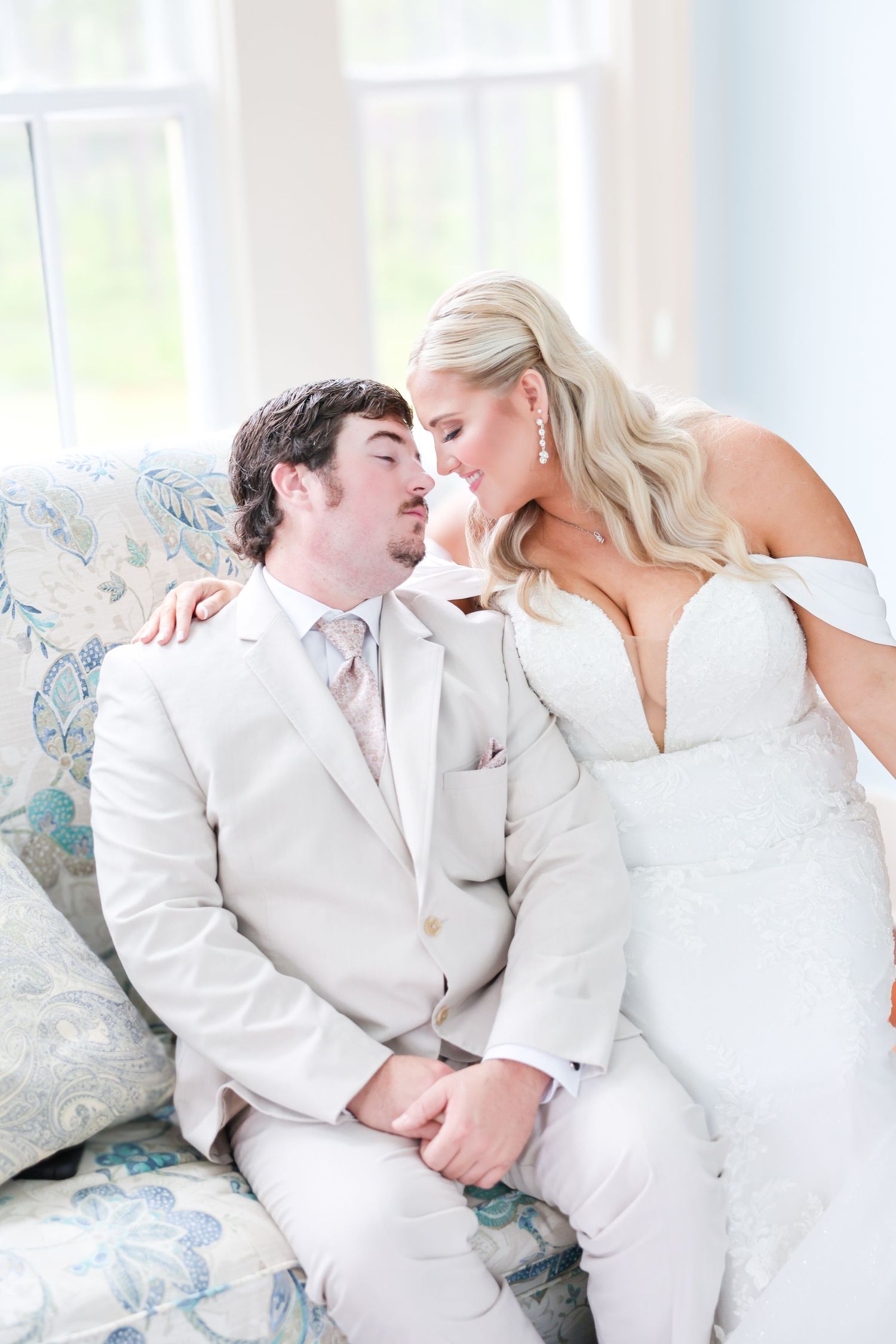 Bride and groom share an intimate moment leaning close on a floral couch in soft natural window light.