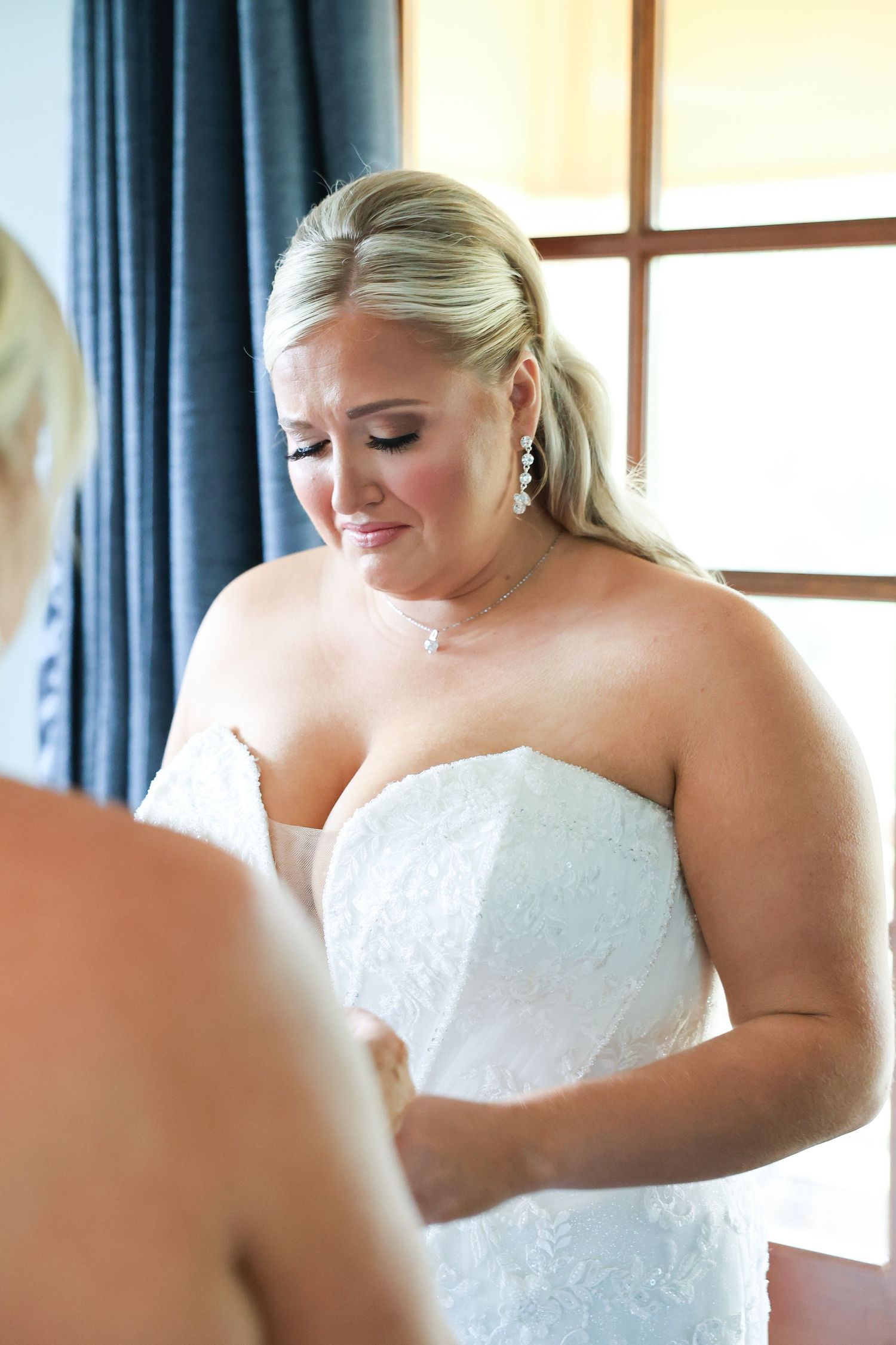 Emotional bride in strapless white lace wedding gown with pearl earrings getting dressed before her ceremony.