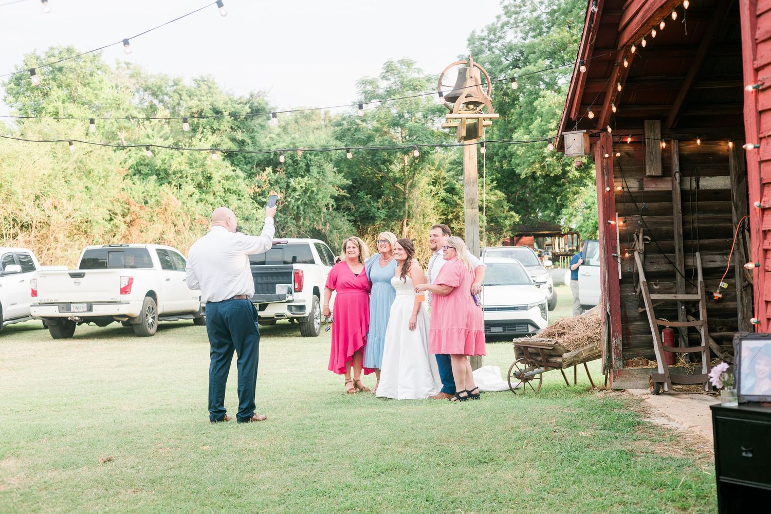 Wedding party gathering outside a rustic red barn venue with string lights and green trees.