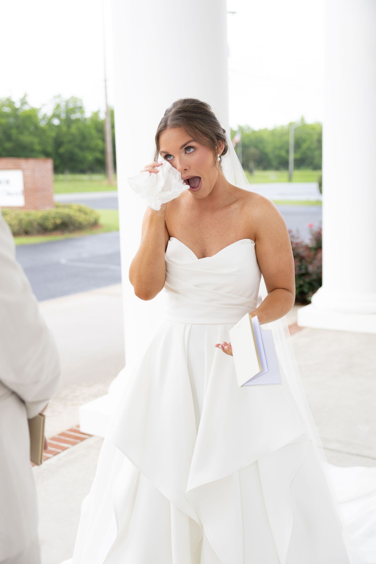 Surprised bride in strapless white wedding dress dabbing eye with tissue while holding letter outdoors.