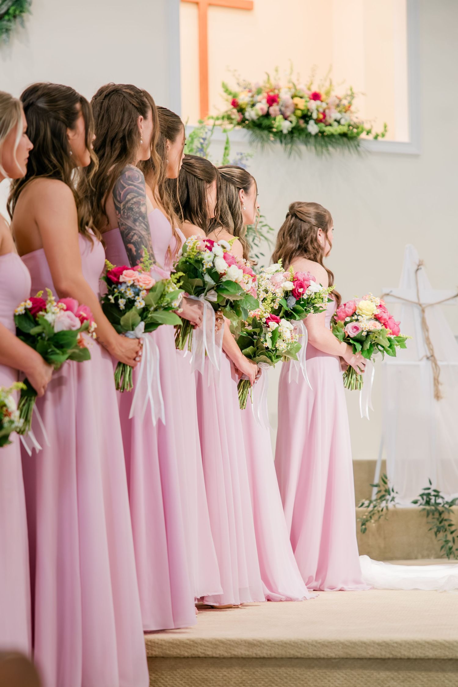 Bridesmaids in pink gowns holding floral bouquets standing at altar during wedding ceremony.