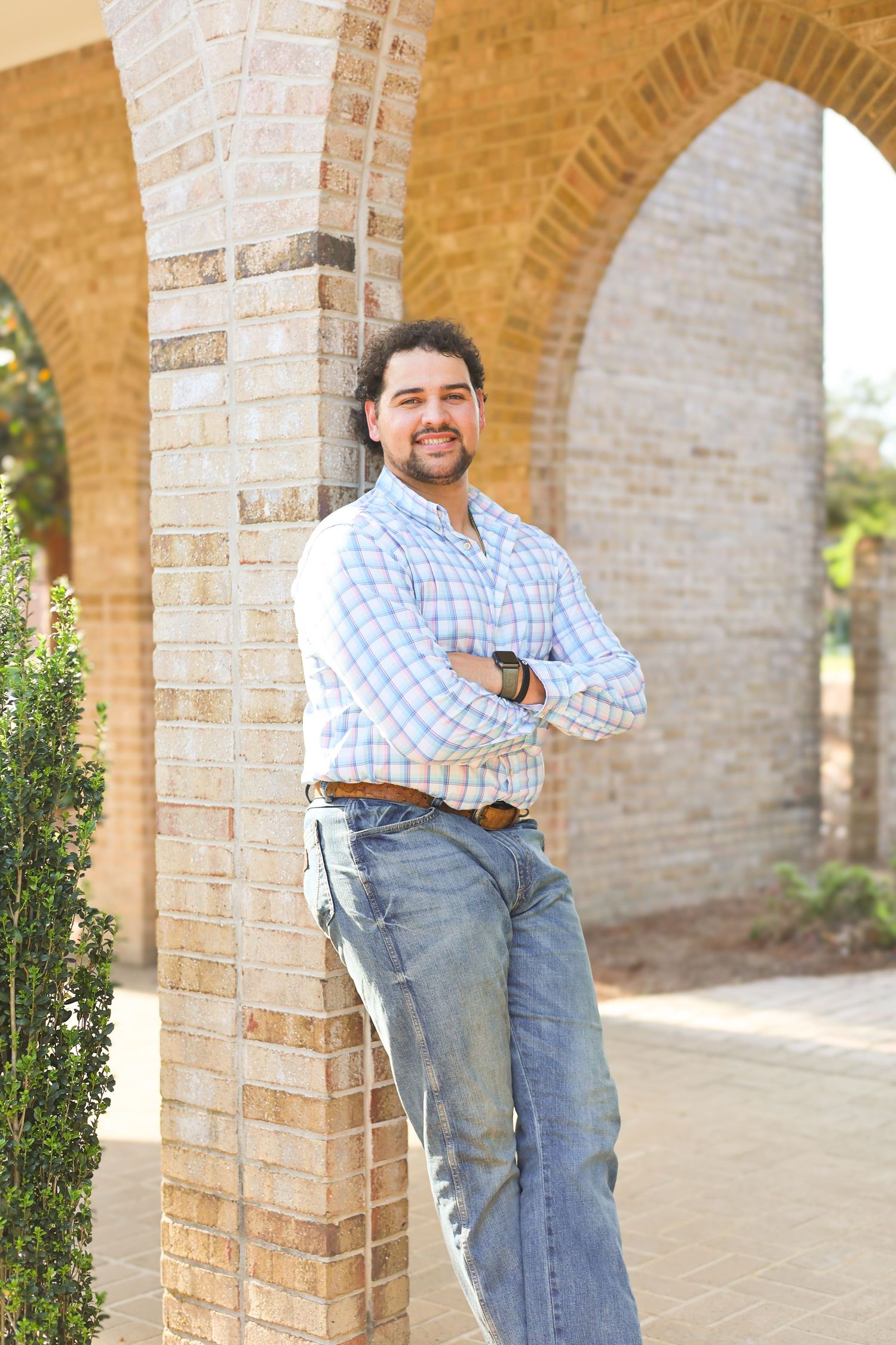Person in checkered shirt leans against brick archway on college campus.
