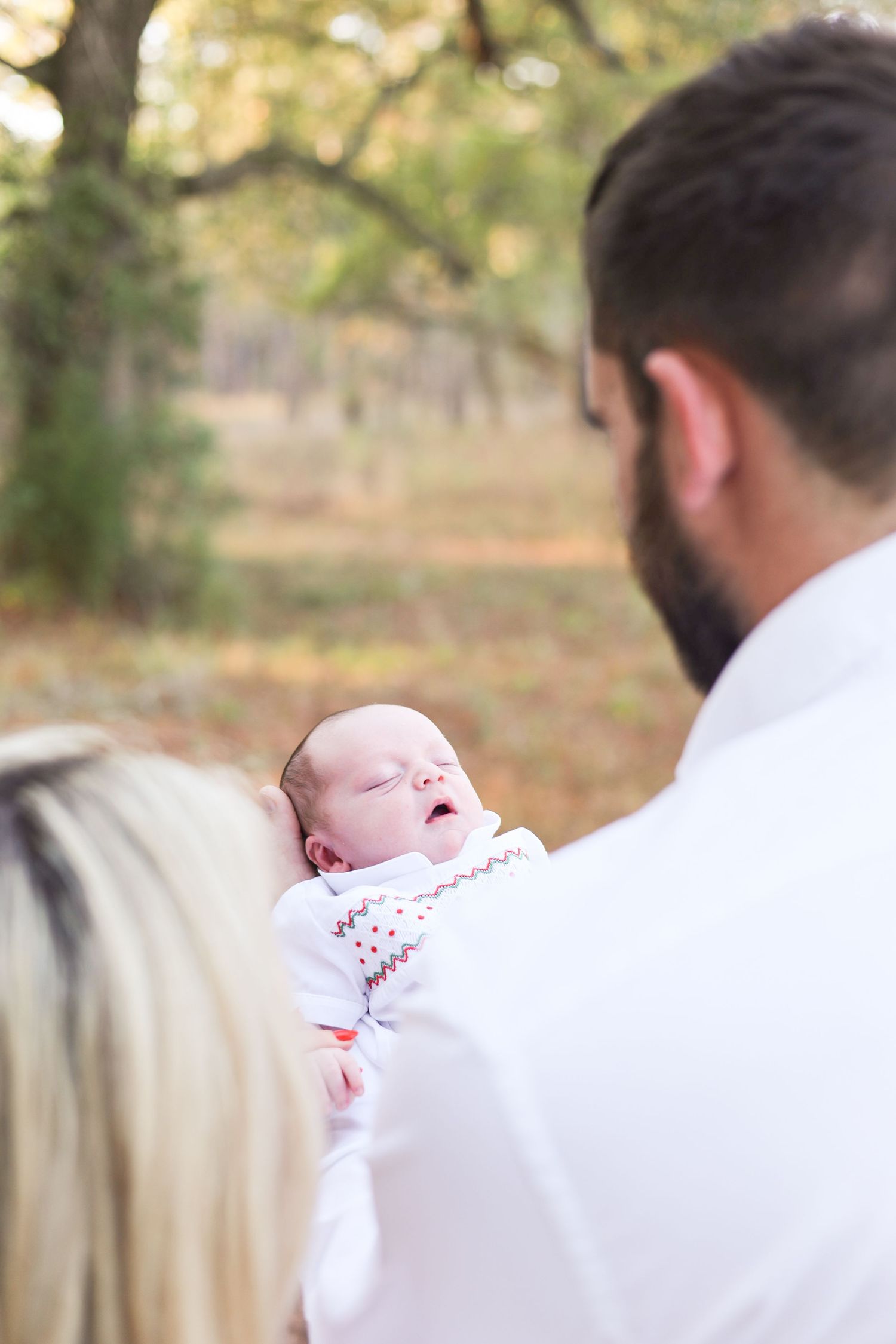 A baby in white clothing being held and laughing during an outdoor photo session with a blurred natural background.