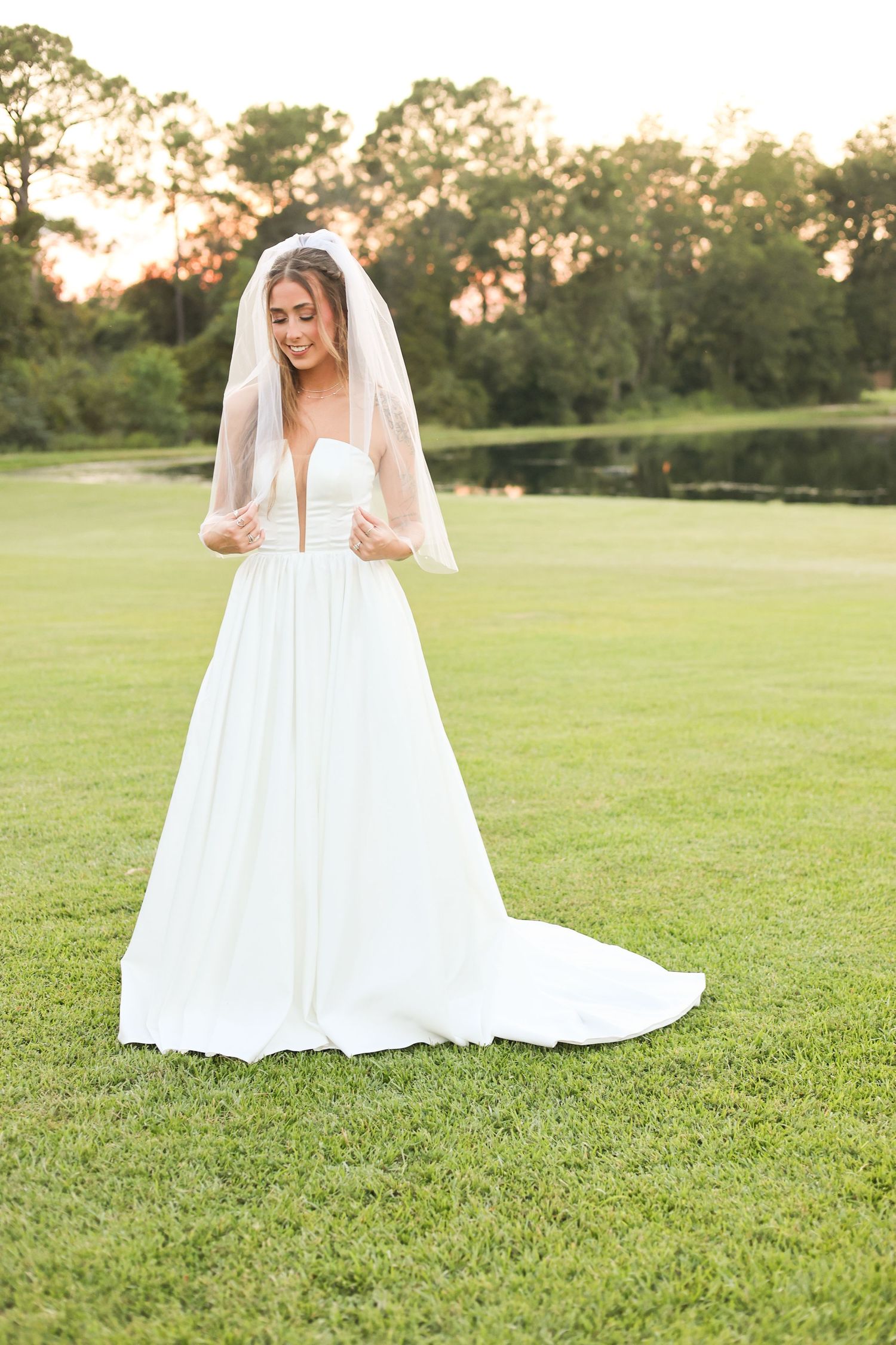 A bride in a plunging neckline gown poses on a sunlit lawn during golden hour.