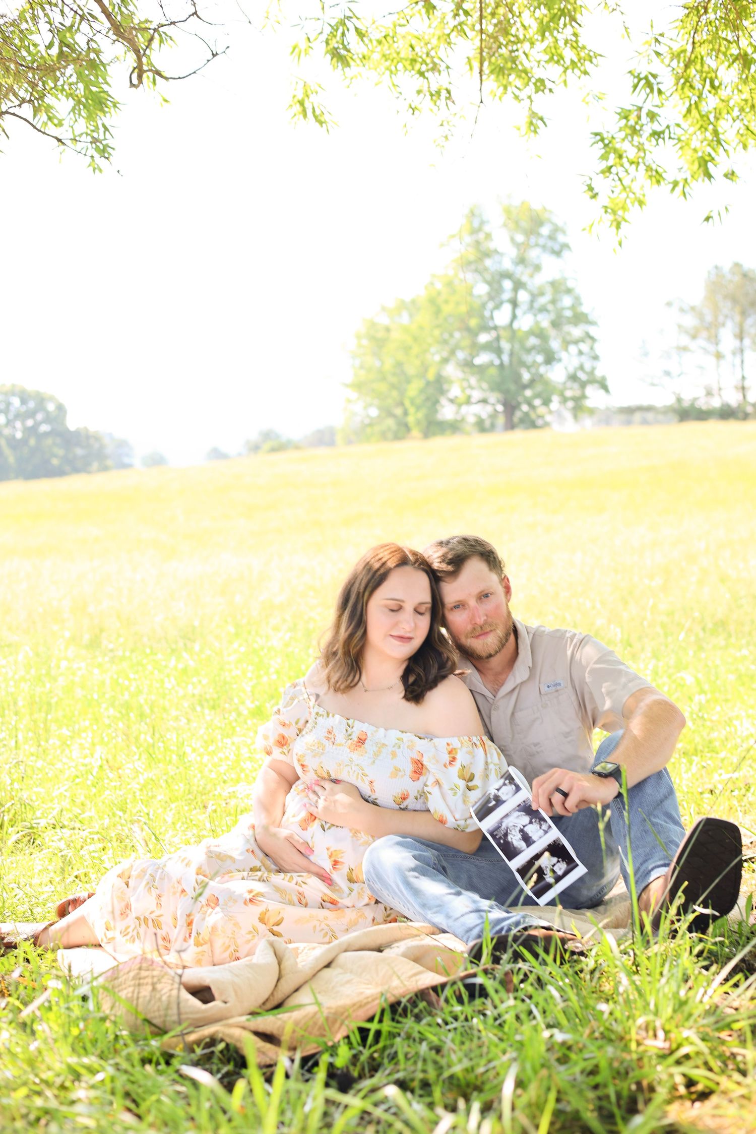 A couple sits in a sunny meadow holding ultrasound photos during a pregnancy announcement photoshoot.