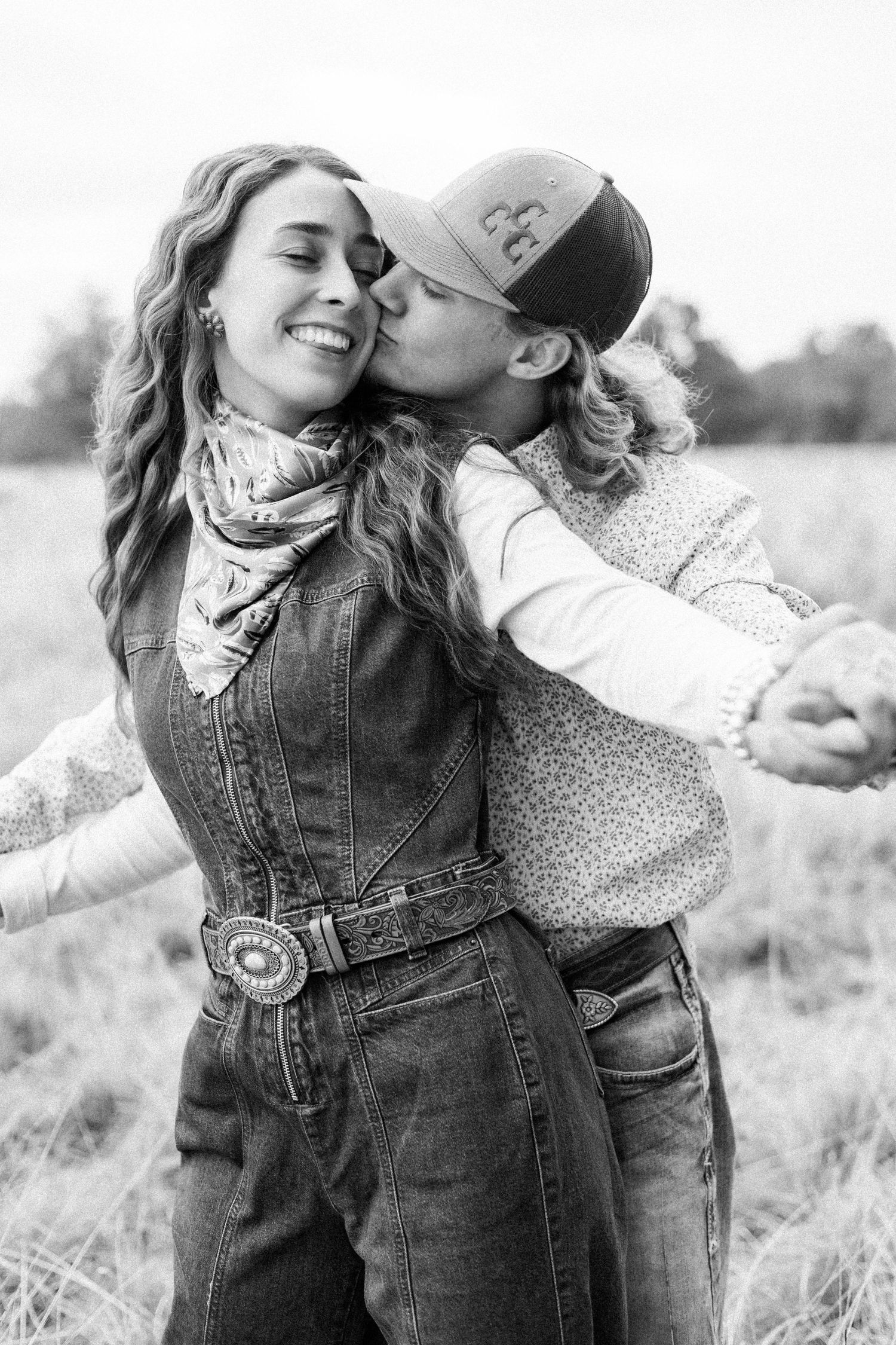 Black and white photo of two women embracing in a field, one kissing the other on the cheek, both smiling.