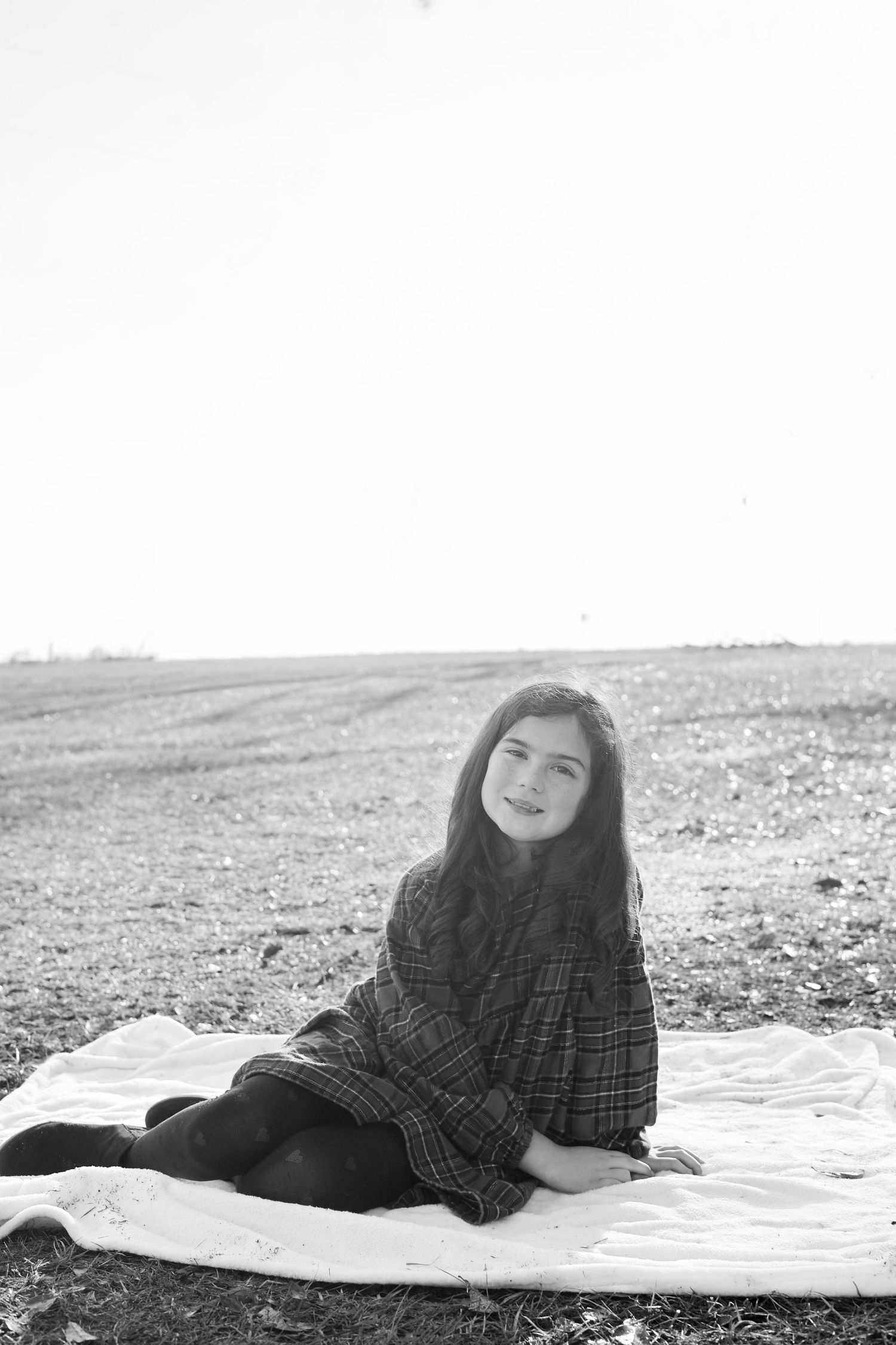 Black and white photo of a young girl sitting on a blanket in an open field, smiling softly at the camera.
