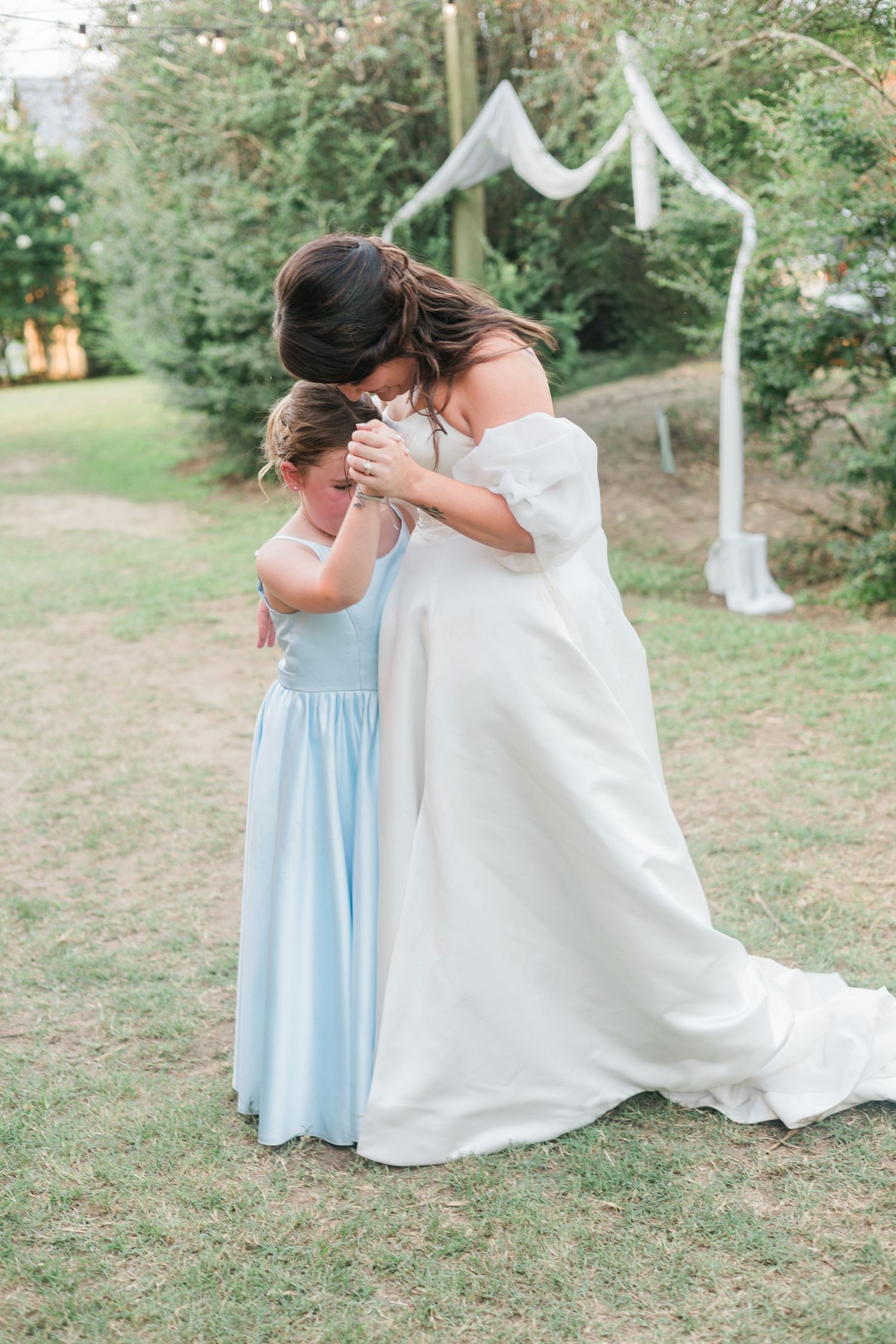 Bride in white off-shoulder gown hugging flower girl in light blue dress at outdoor wedding ceremony.