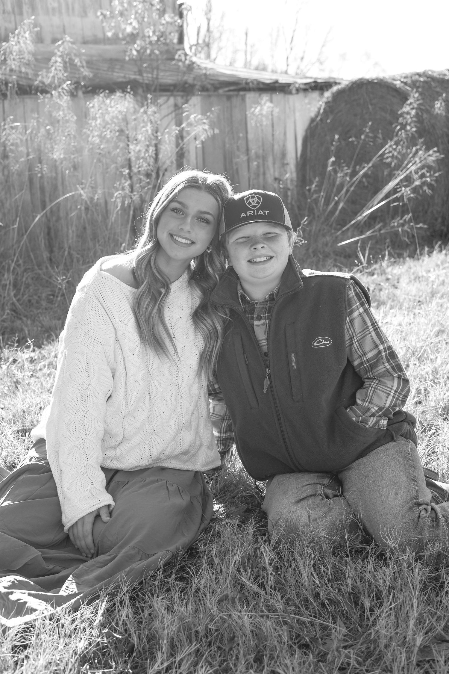 Black and white portrait of a couple relaxing in tall grass by a weathered wooden fence.