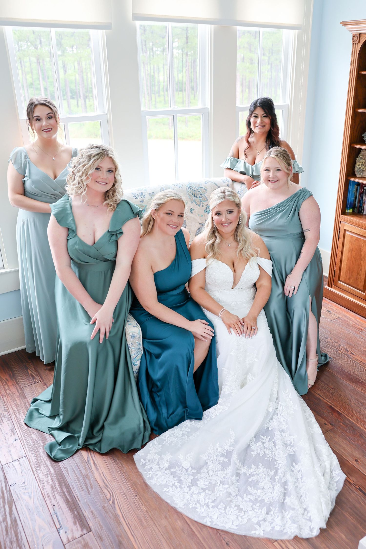 Bride in white lace gown poses with five bridesmaids in sage and teal satin dresses indoors.