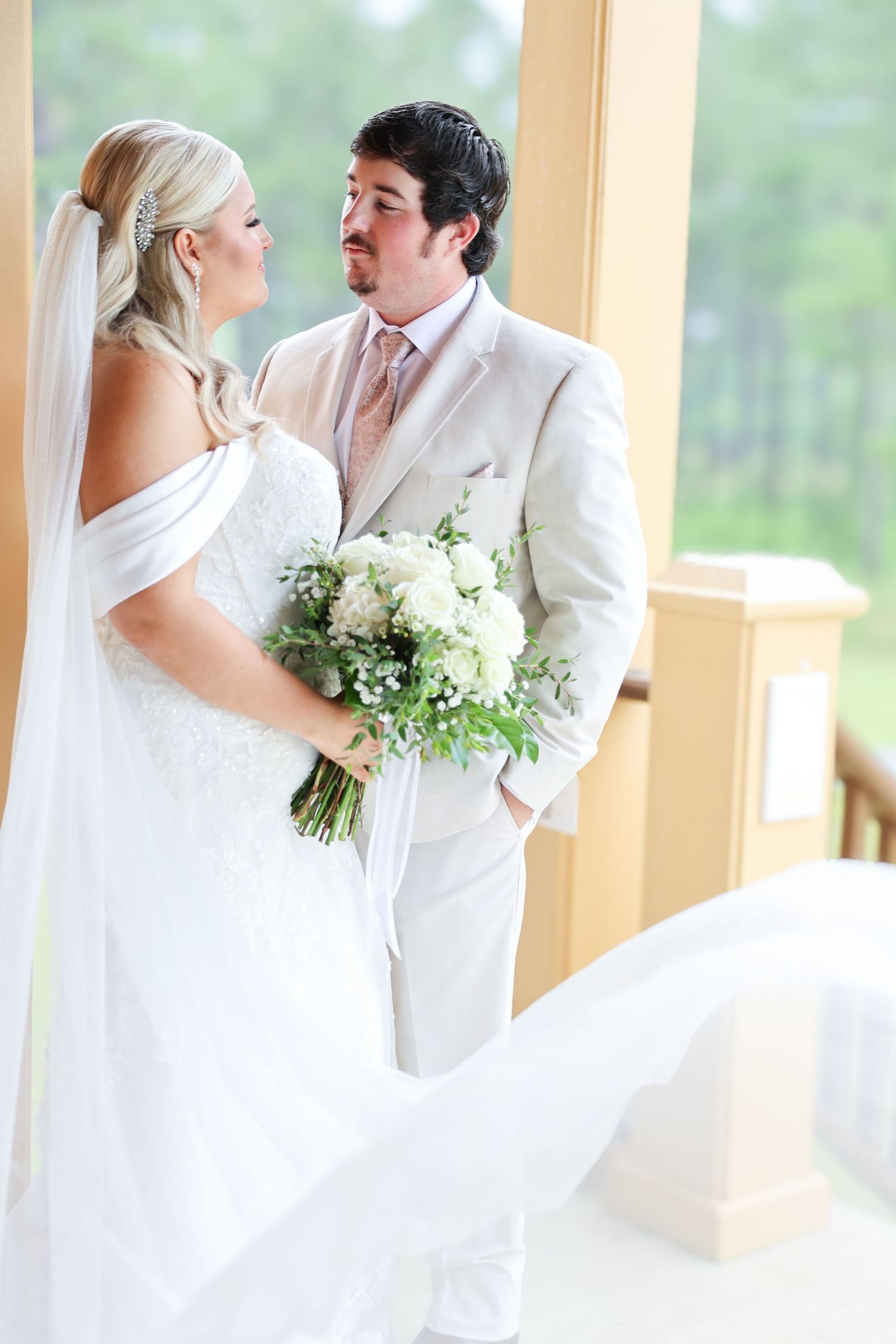 Bride in white gown holding bouquet gazes at groom in tan suit near window at elegant wedding venue.