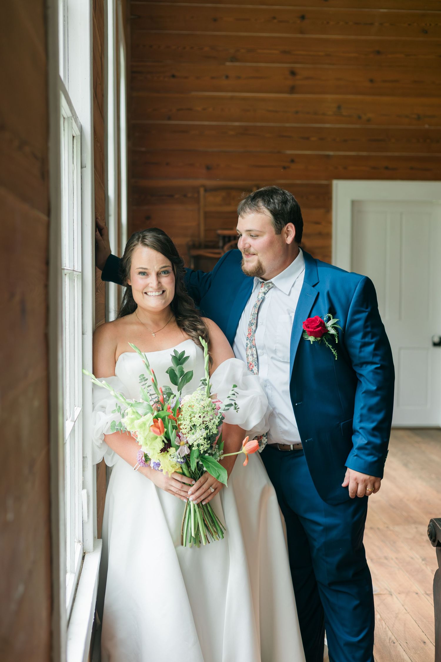 Bride in off-shoulder gown holding wildflower bouquet with groom in blue suit by window.