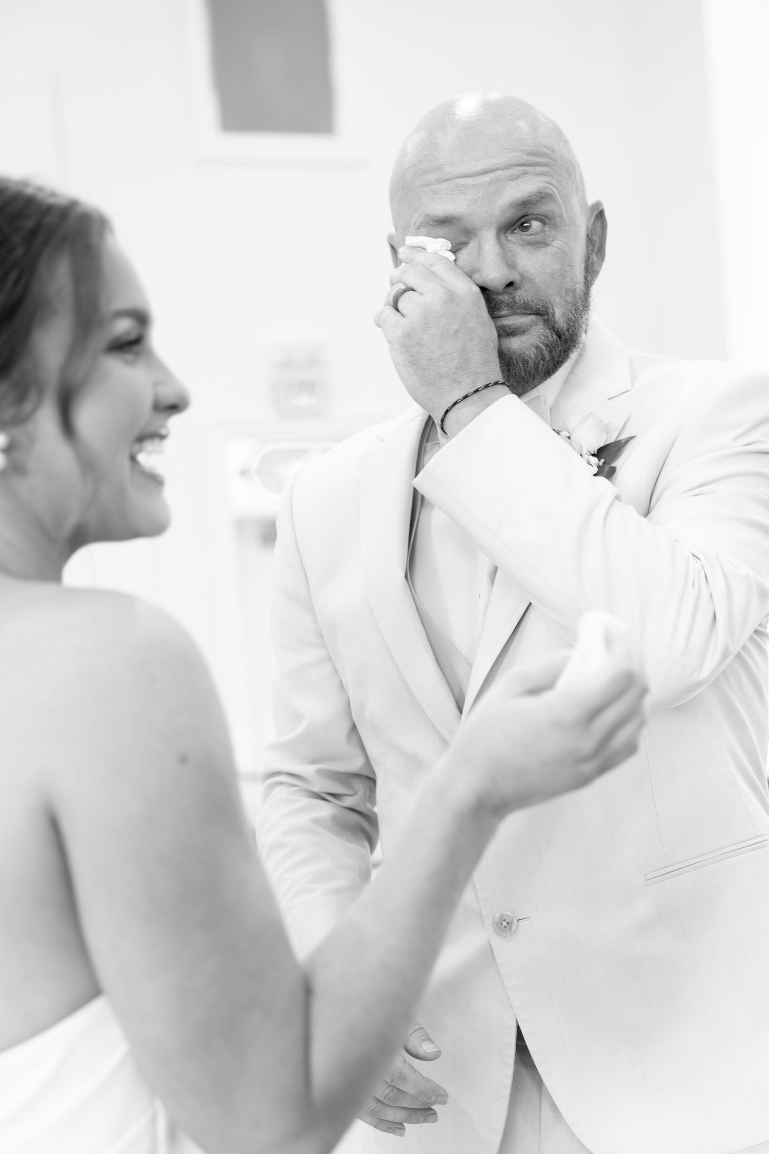 Emotional groom in white suit wipes tears while holding brides hands in black and white wedding photo.