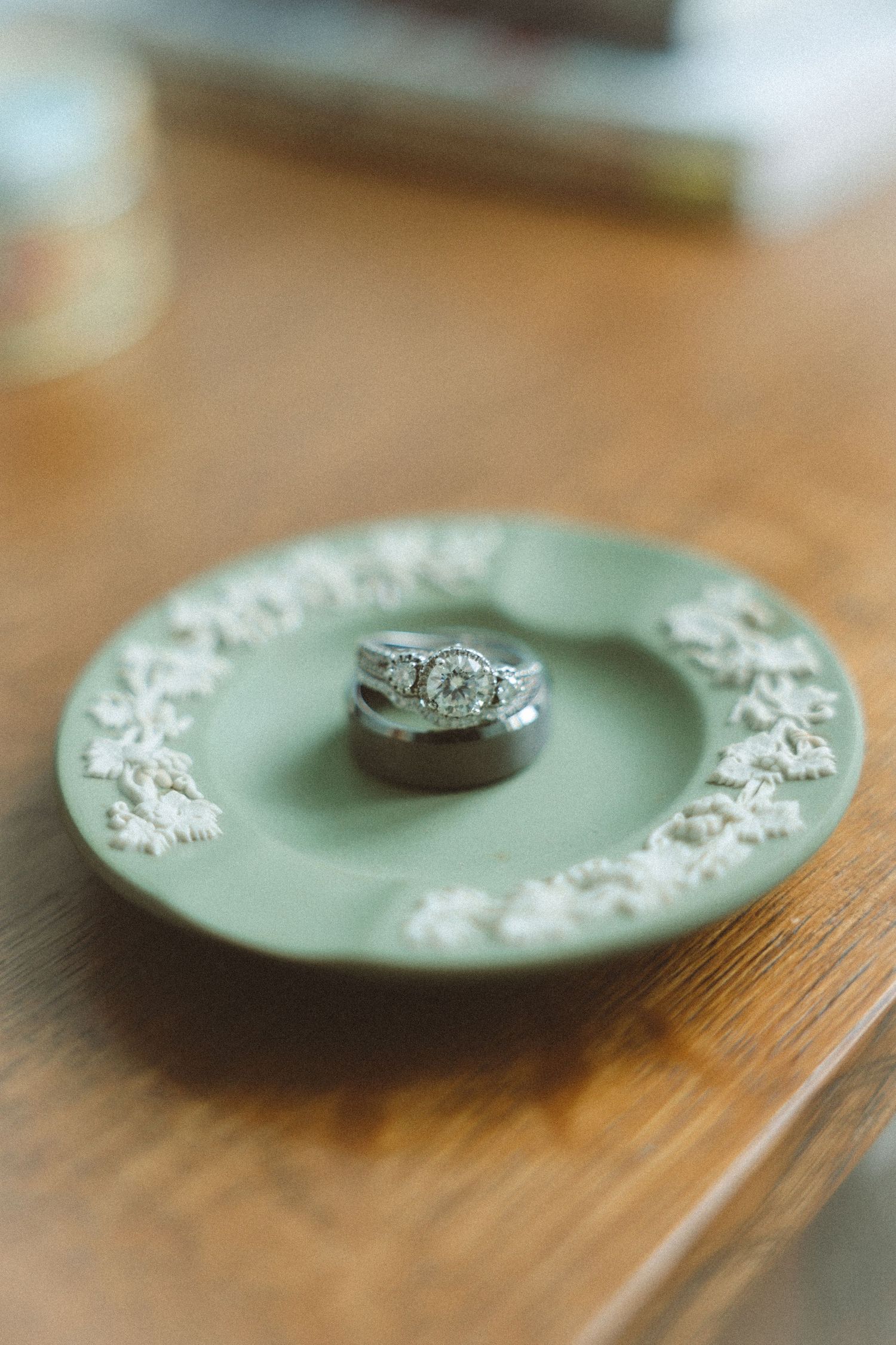 Silver diamond engagement ring and wedding band displayed on a green Wedgwood decorative plate on a wooden surface.