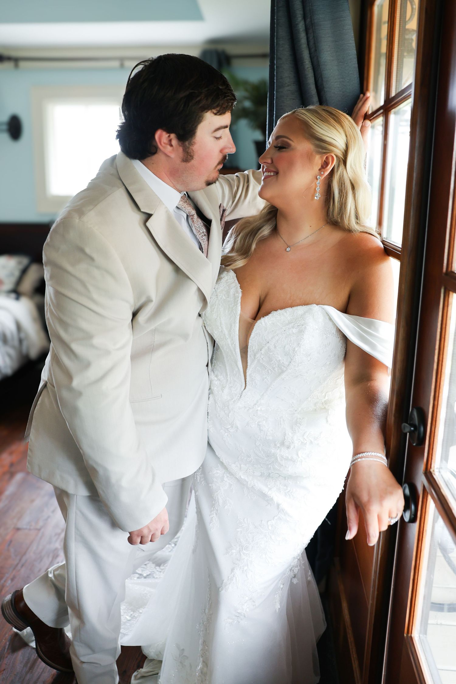 Bride and groom sharing a romantic moment by a window, she in a white off-shoulder gown, he in a tan suit.