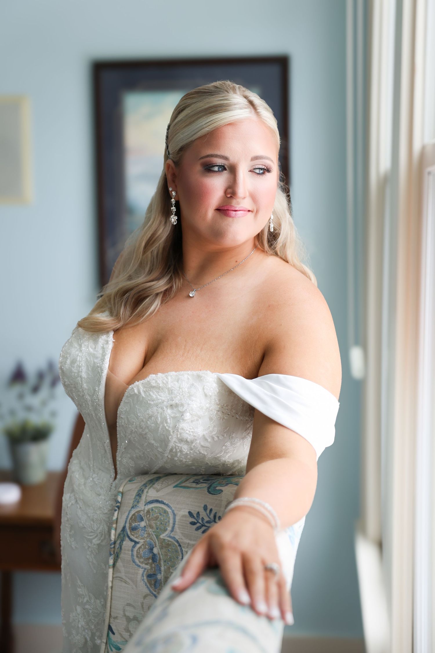 Elegant bride in off-shoulder lace wedding gown with pearl earrings and necklace posing by a window.