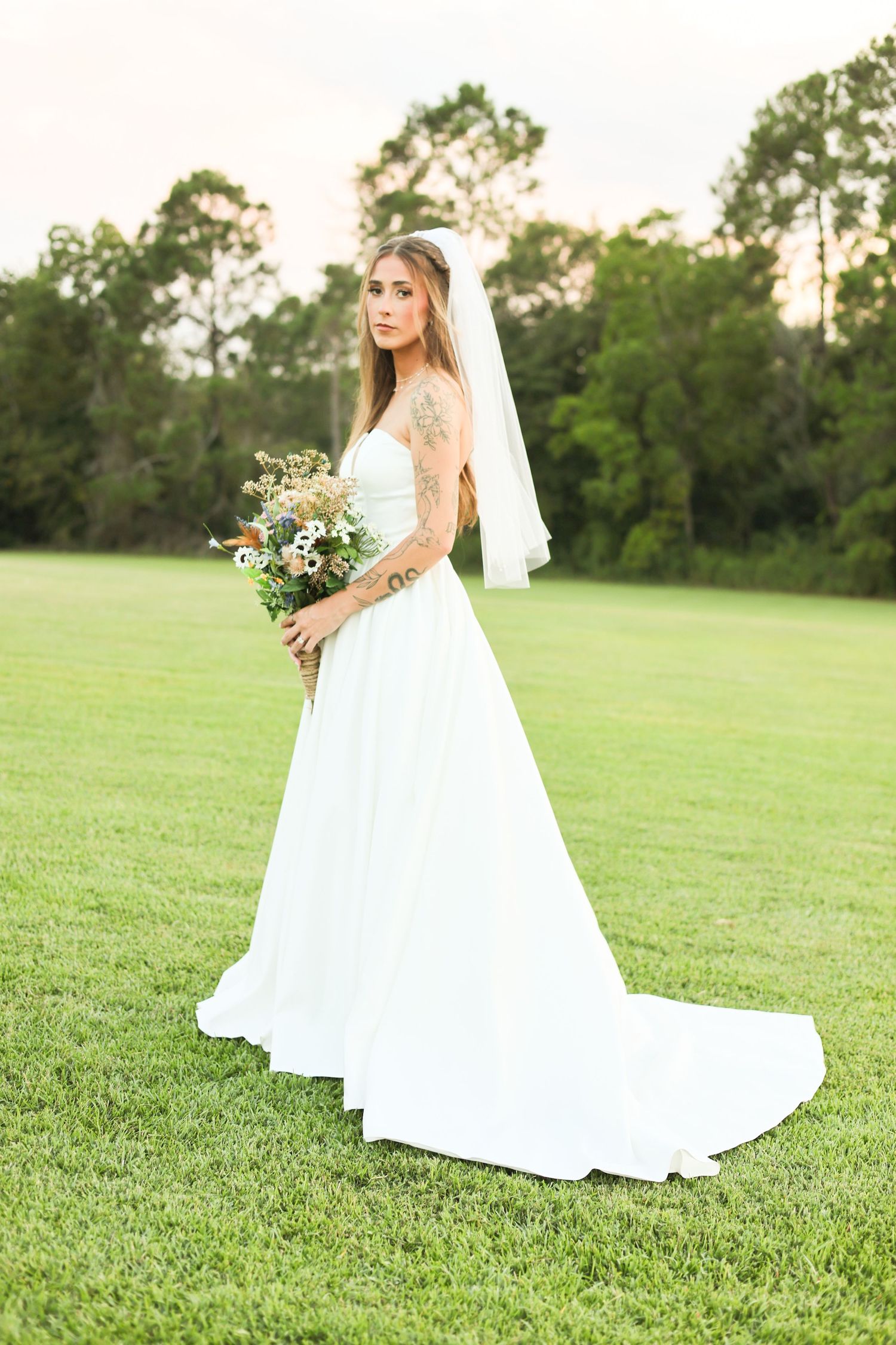 A bride holds a wildflower bouquet while standing in an open field at golden hour.