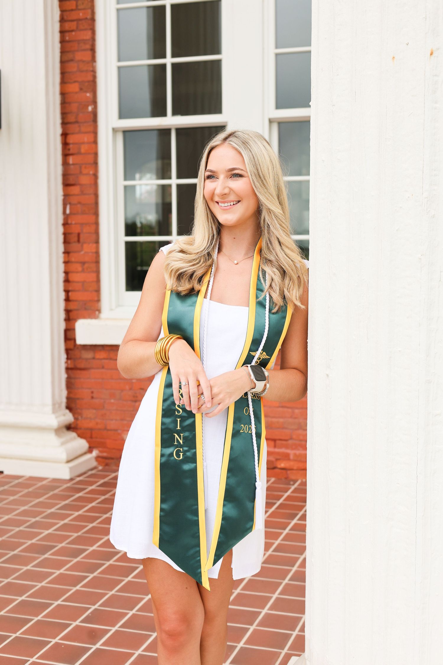 Graduate wearing green sash and white dress poses in front of brick building.