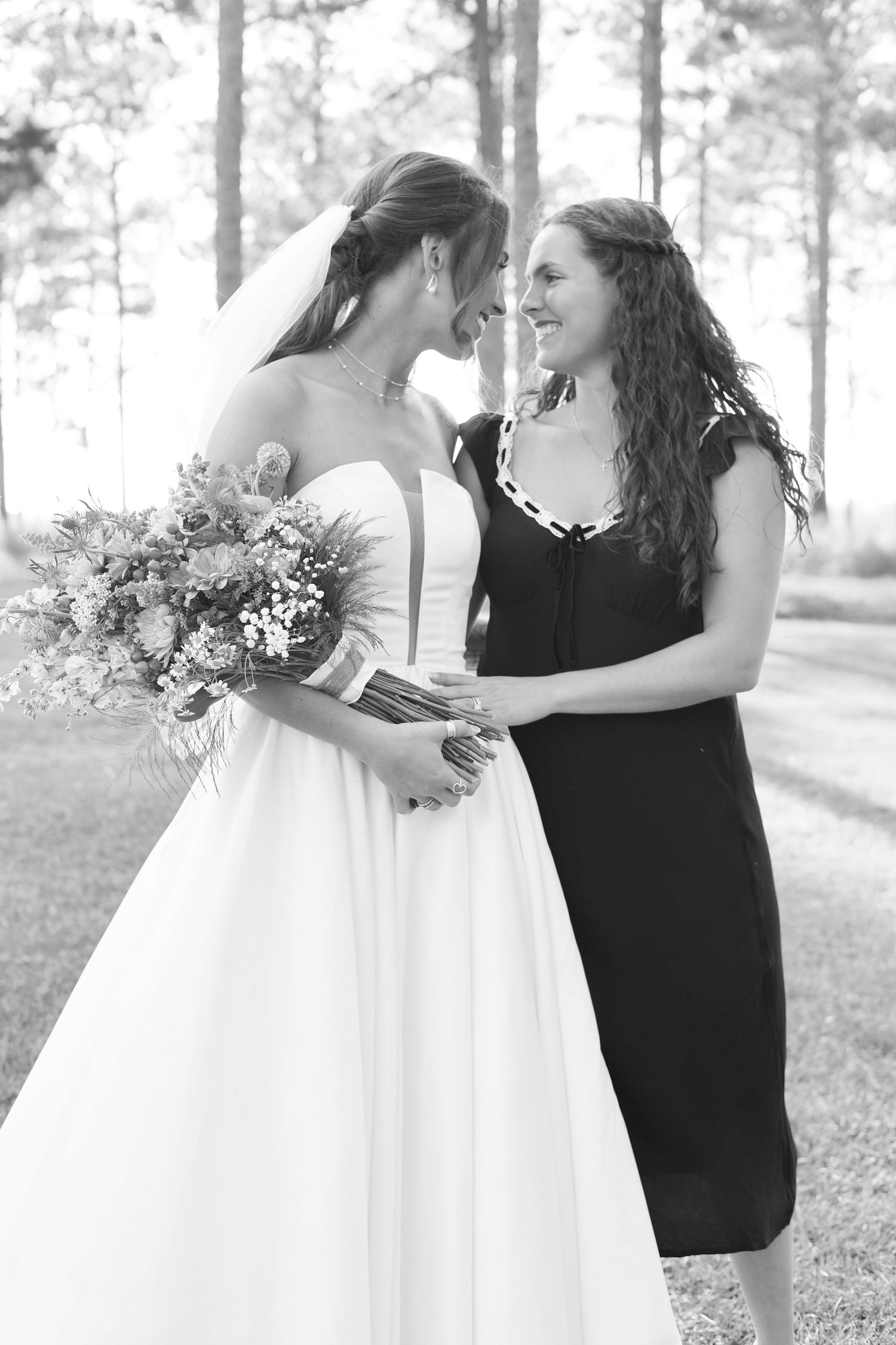 A bride in a white gown shares a tender moment with someone in a black dress among tall pine trees.