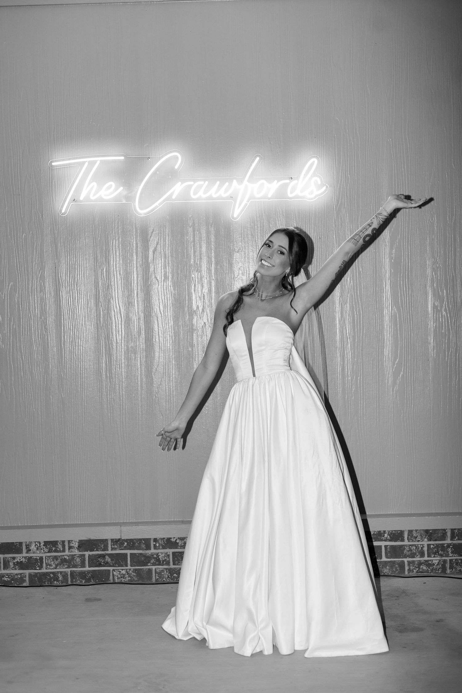 Bride in strapless ball gown poses joyfully in front of The Crawfords neon sign in black and white wedding photo.