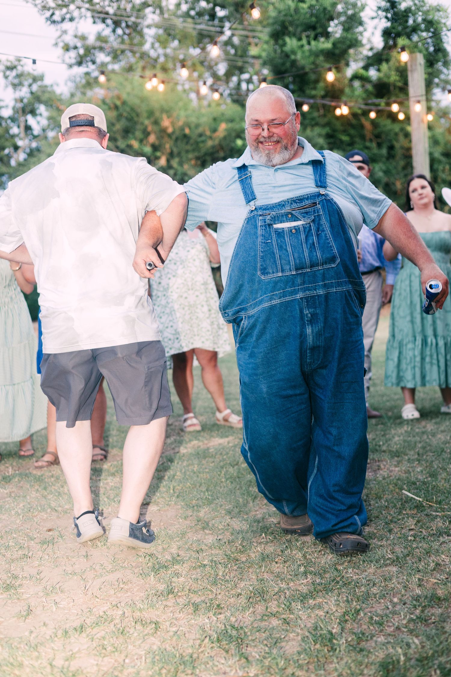 Cheerful man in denim overalls dancing with guests at an outdoor wedding reception.