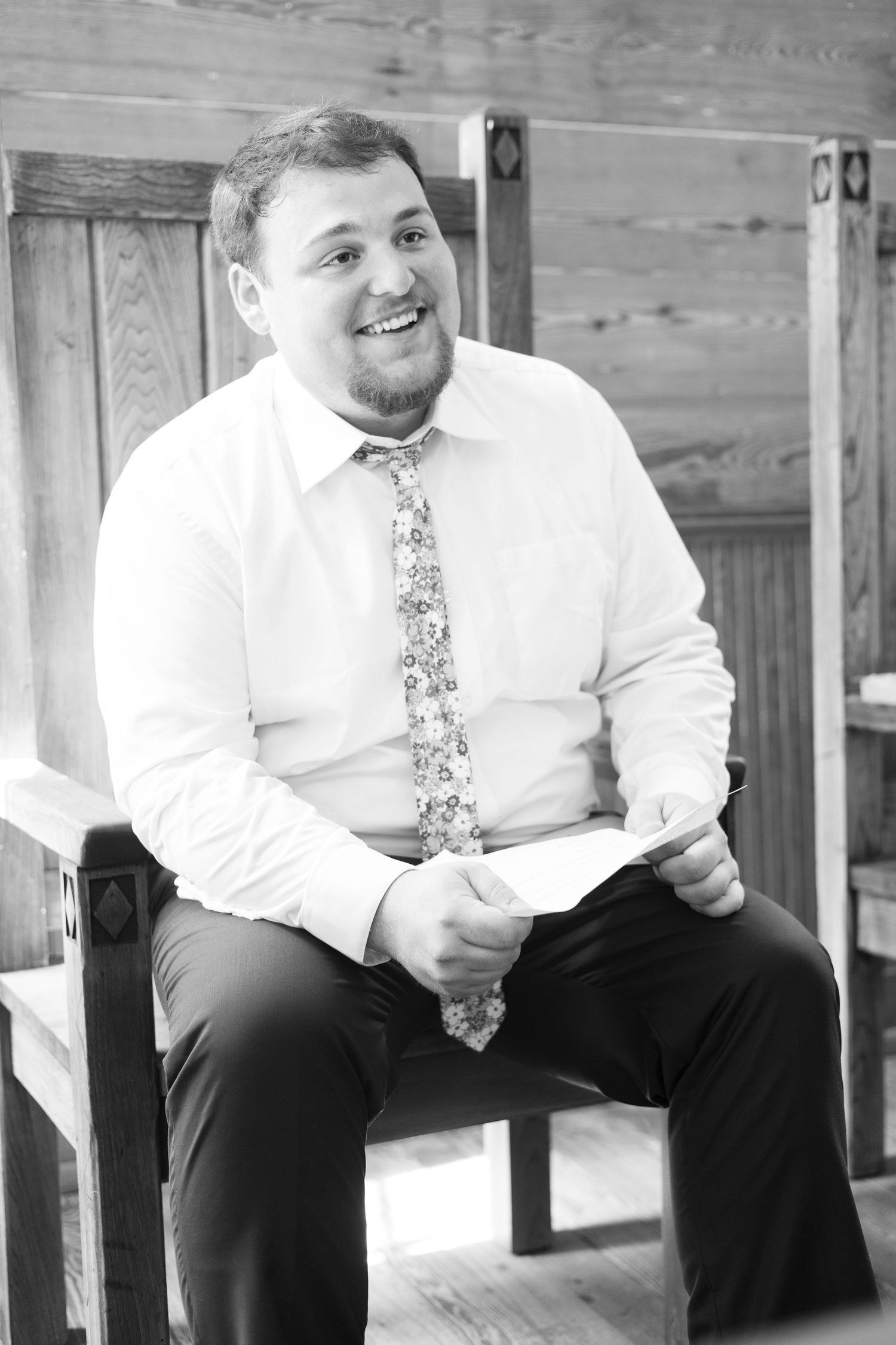 Smiling groom in white dress shirt and floral tie sits on wooden chair holding vows card in black and white portrait.