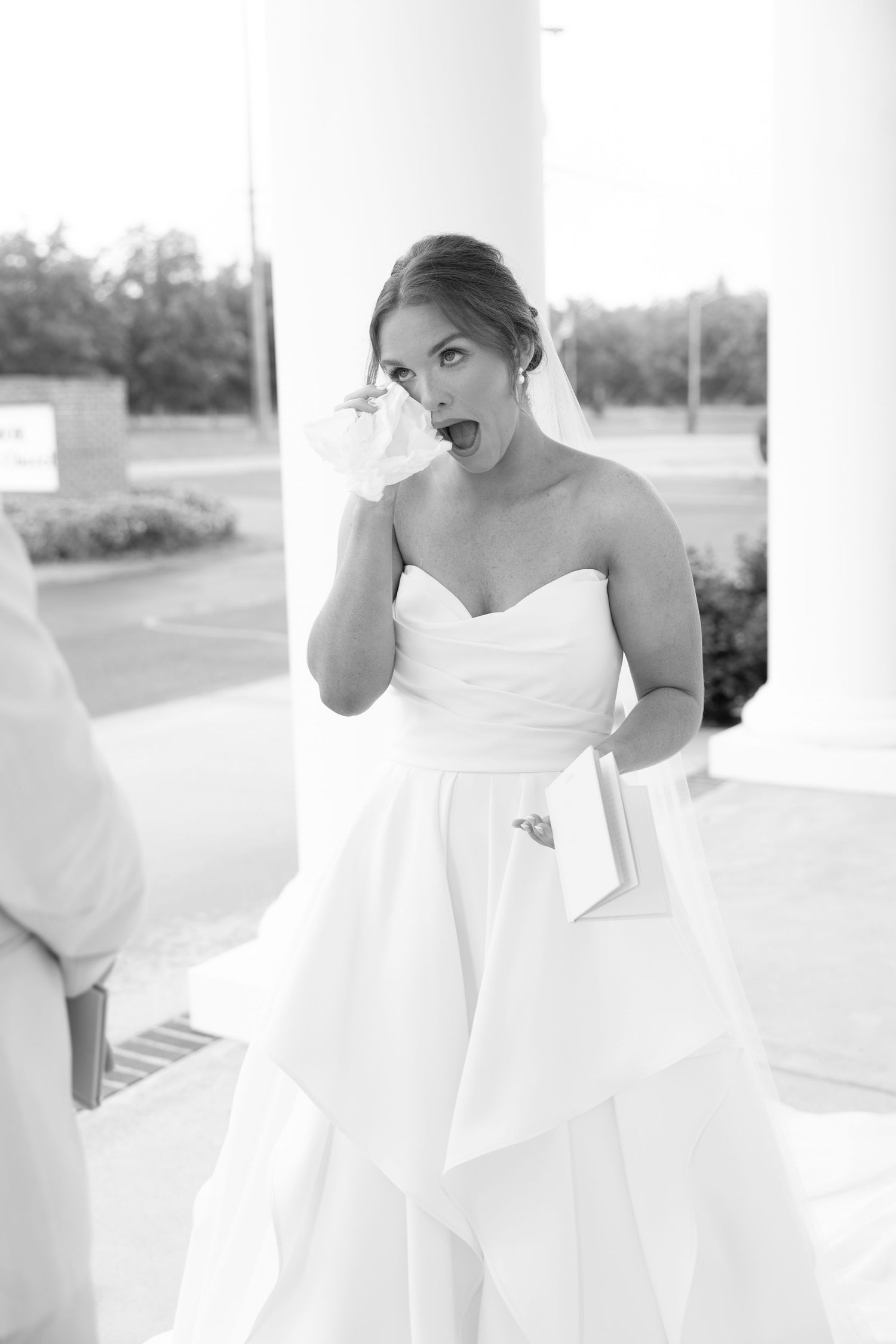 Emotional bride in strapless white gown dabbing tears with handkerchief outside church on wedding day.