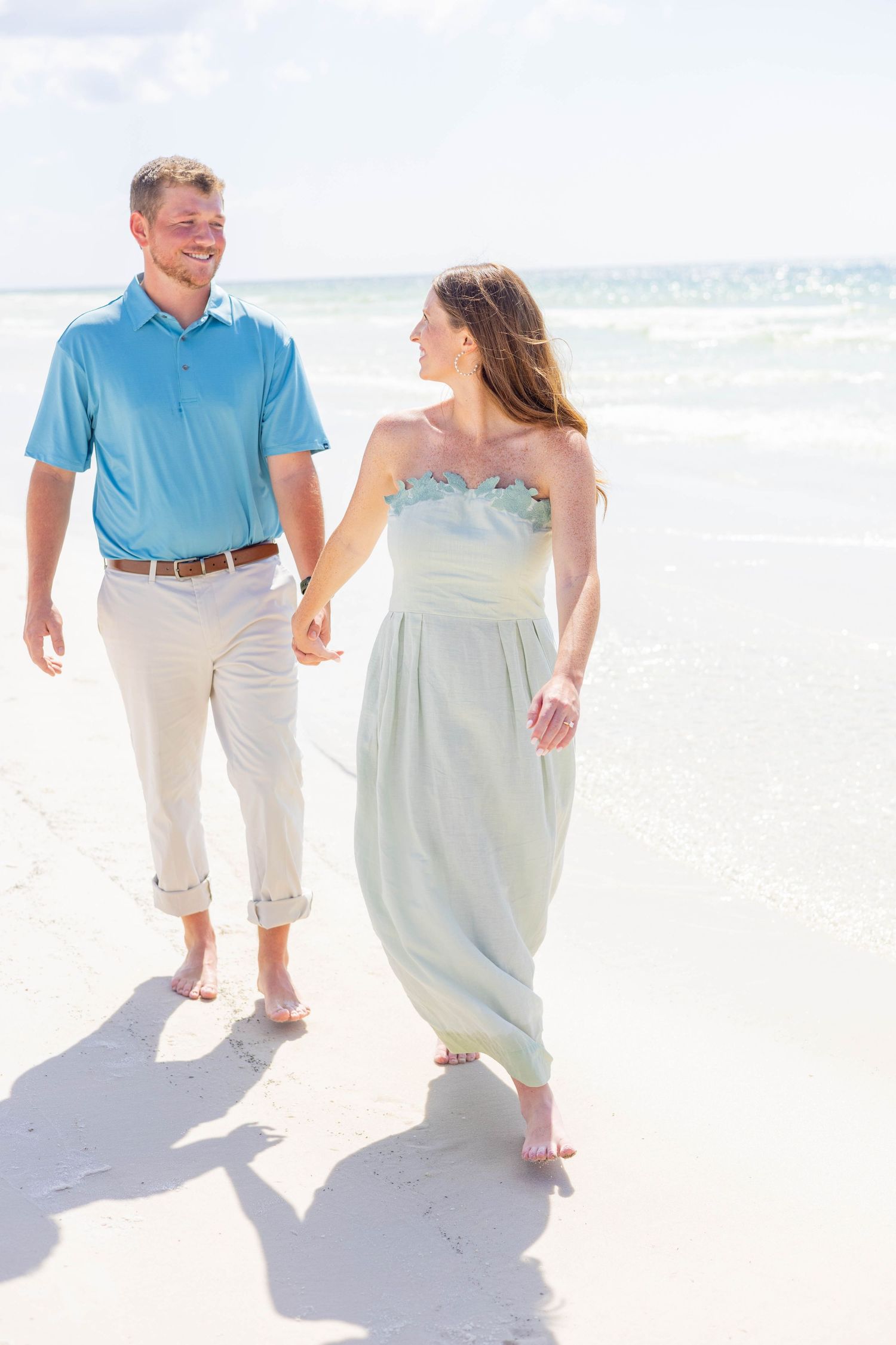 Joyful couple holding hands and walking barefoot on white sand beach, man in blue polo and woman in mint dress.