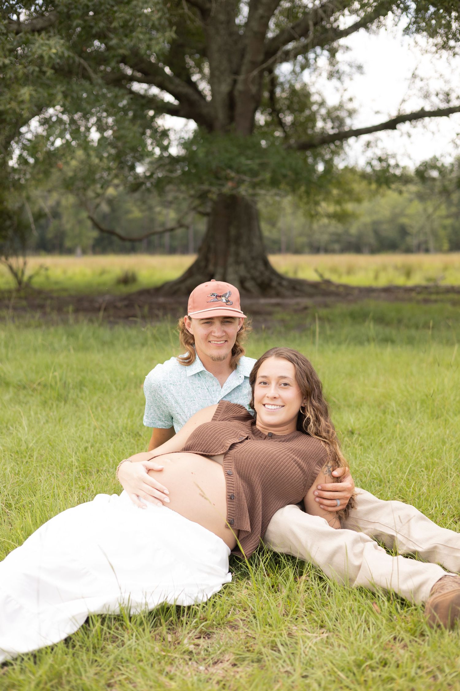 Couple sitting in green field, pregnant woman leaning on partner near large oak tree outdoors.