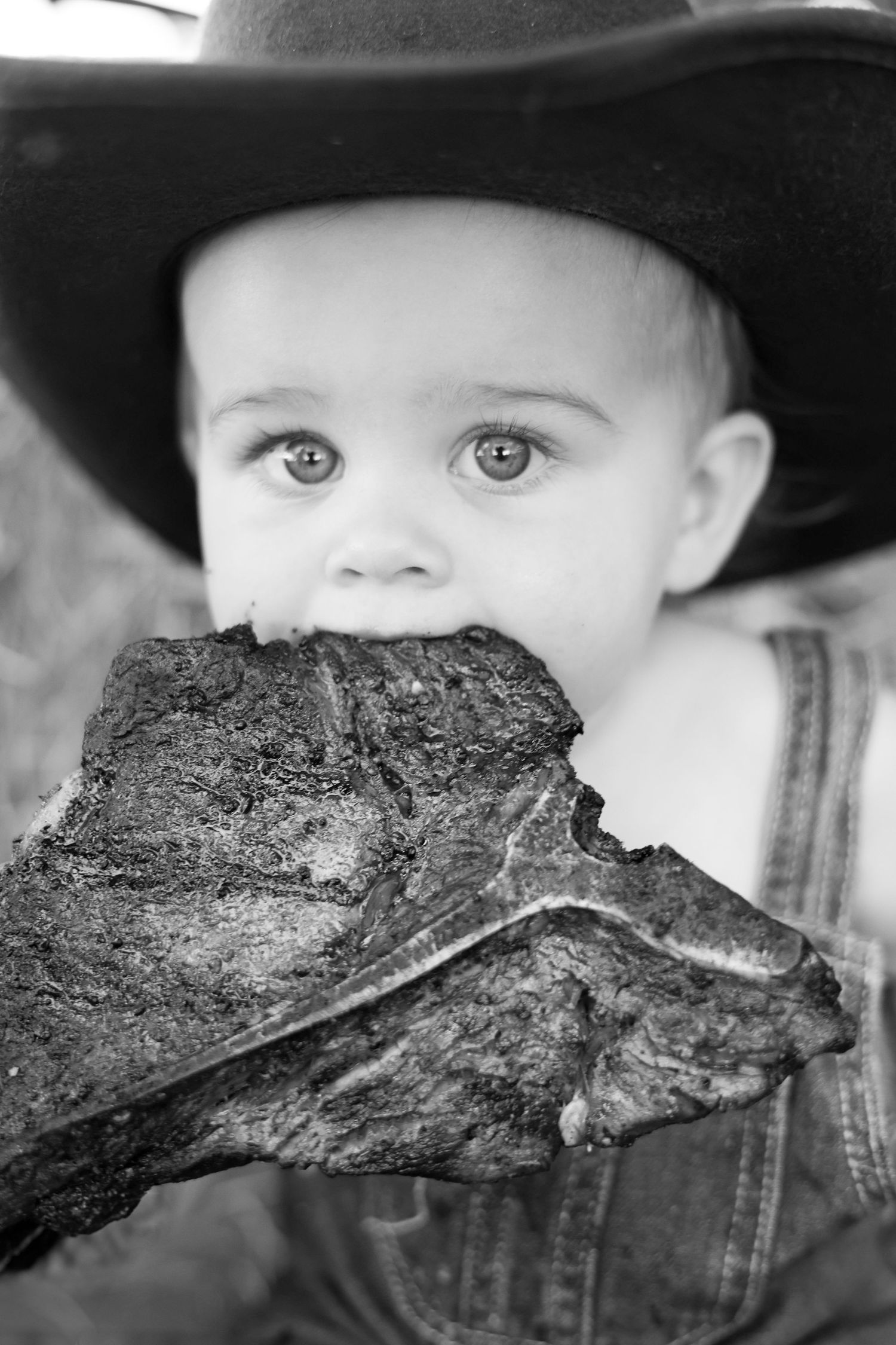 Black and white photo of a baby in a cowboy hat biting into a large grilled steak.