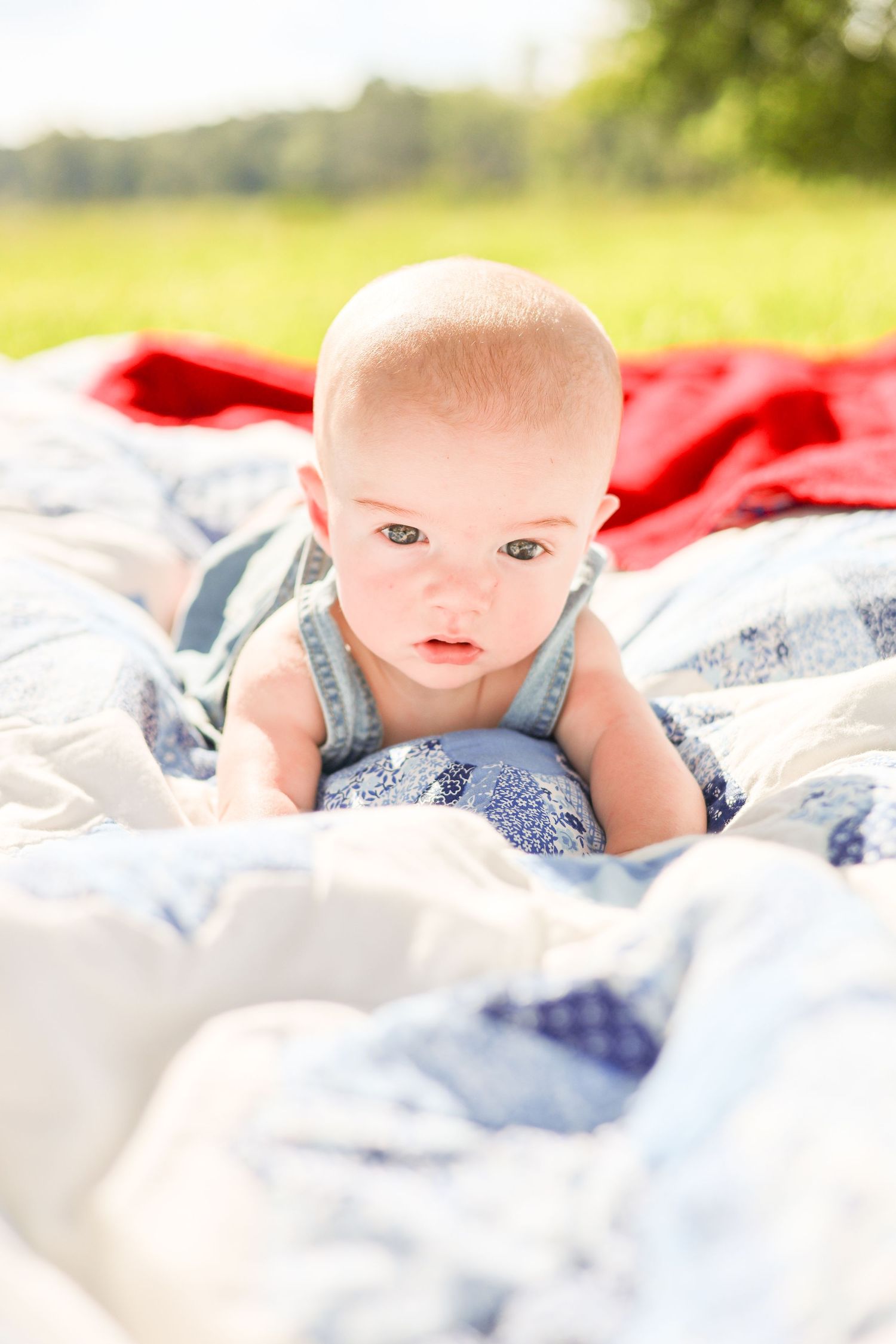 Curious baby in blue outfit doing tummy time on a patchwork quilt outdoors on a sunny day.