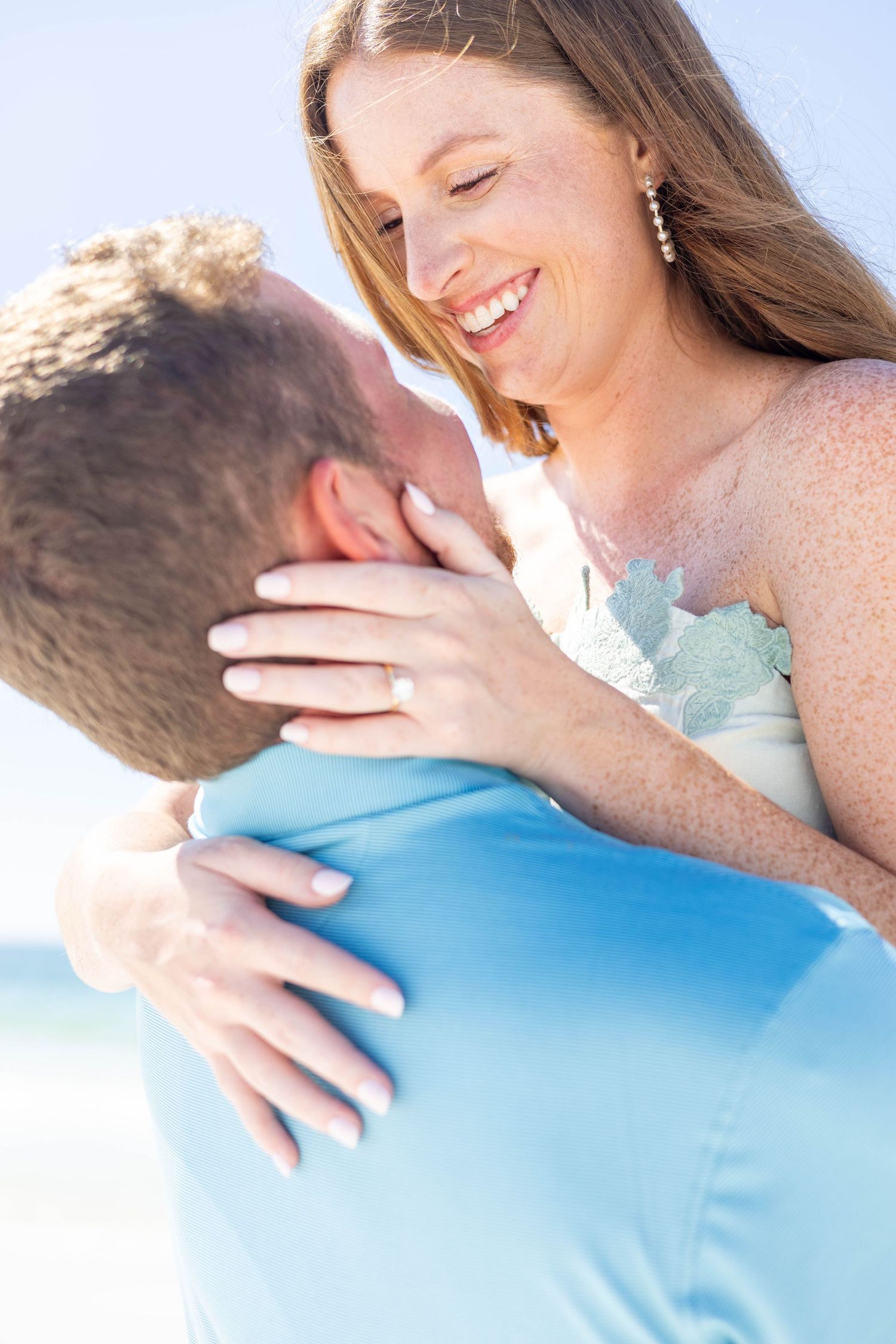 Happy bride with freckles laughing while being lifted by groom in blue shirt during romantic beach photo session.