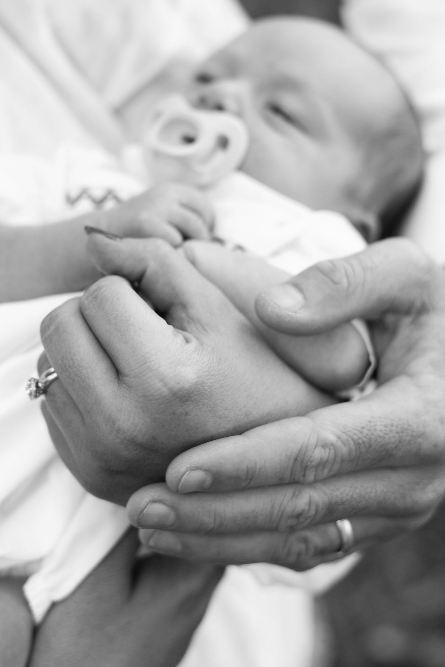 Close-up black and white photo of gentle hands holding a sleeping newborn baby's tiny hand.