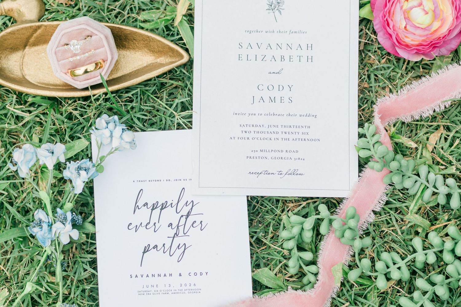 Elegant wedding invitation suite with calligraphy script on grass, featuring rings in gold dish and pink floral accents.