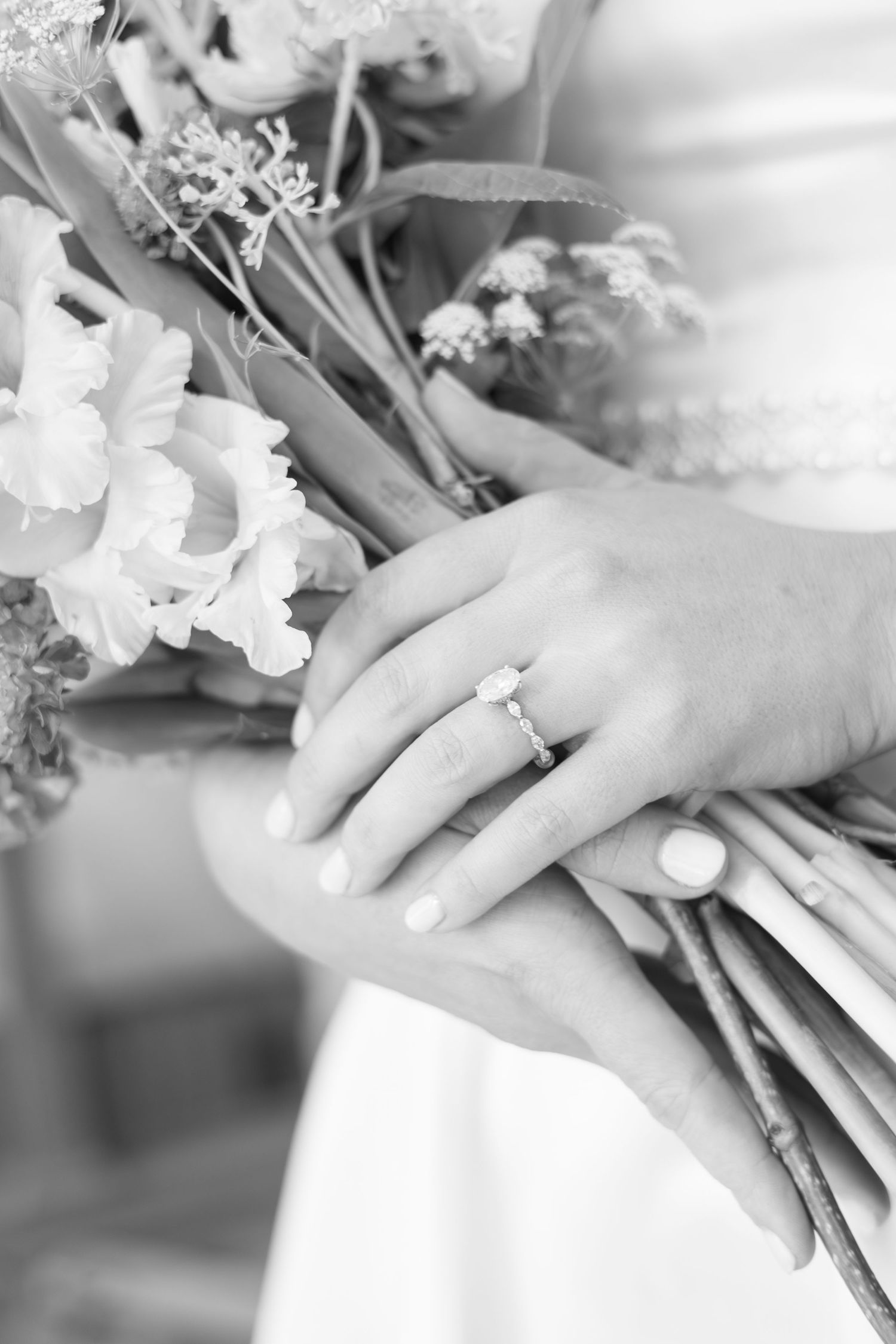 Close-up of bride's hands holding wedding bouquet with diamond engagement ring in black and white.