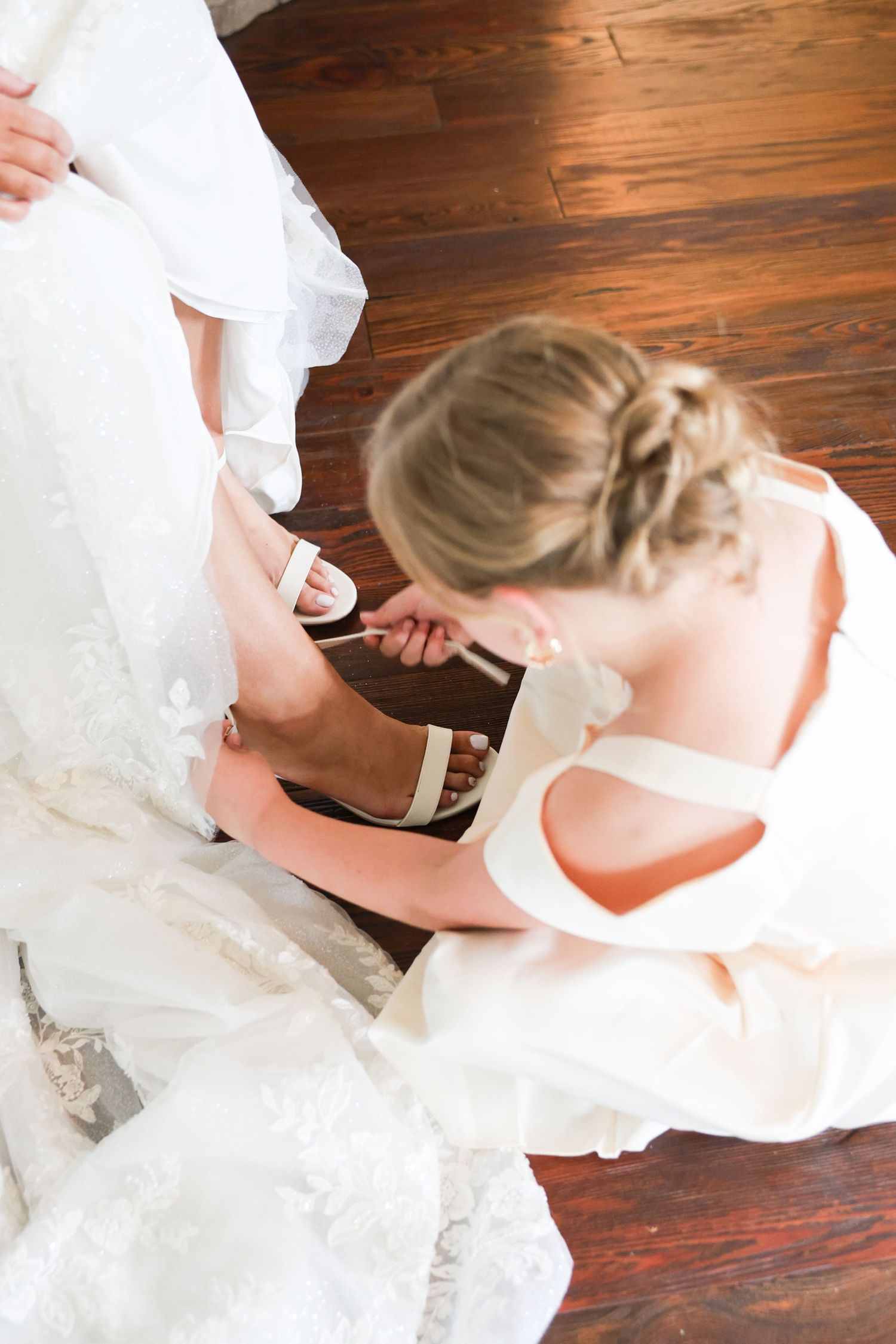 Bridesmaid helping blonde bride put on white strappy wedding sandals on hardwood floor.