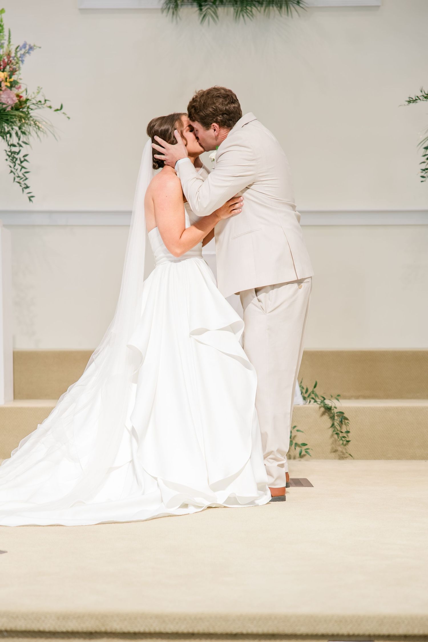 Bride and groom share their first kiss at the altar during a wedding ceremony, bride wearing a white gown with veil.