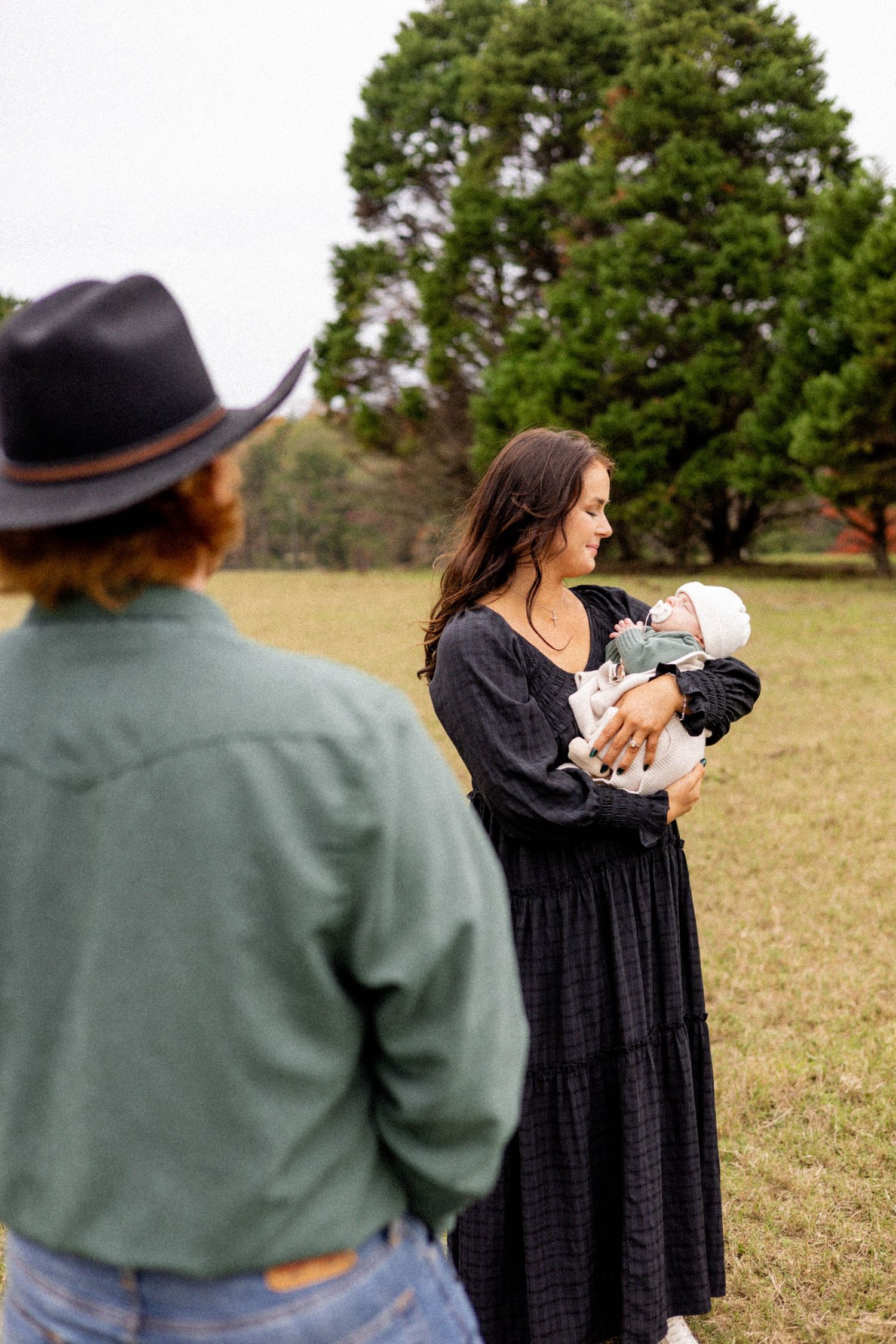 A couple shares a tender moment while holding a baby in an outdoor field setting.