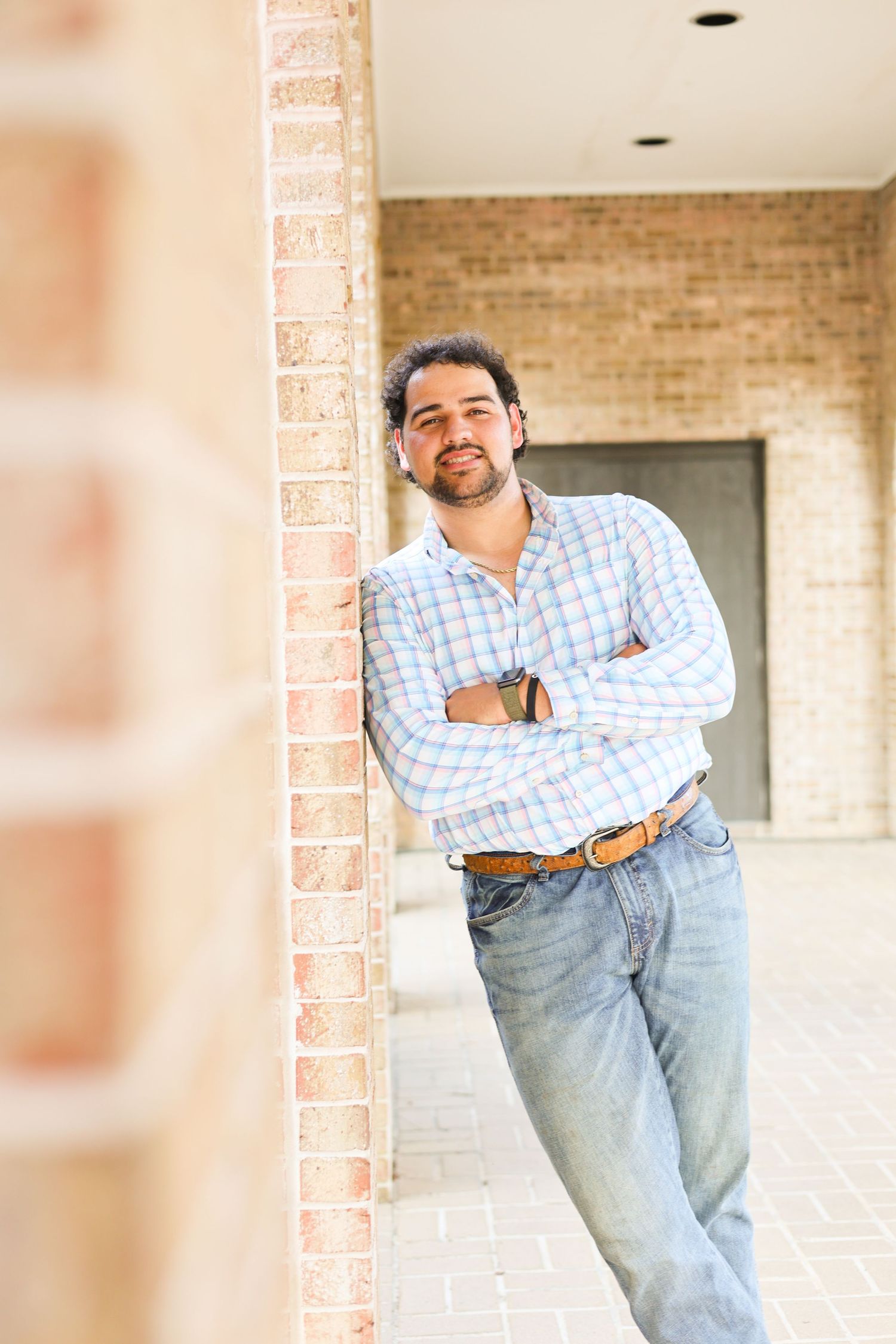 Person in blue checkered shirt poses against brick wall in casual business attire.