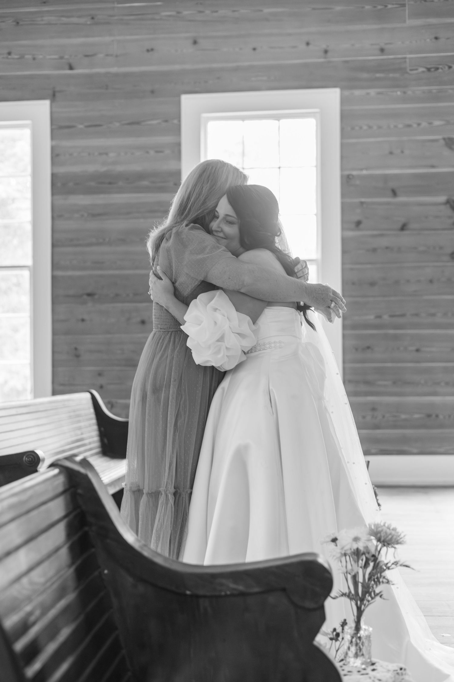 Black and white photo of bride sharing emotional hug with bridesmaid inside rustic wooden chapel.
