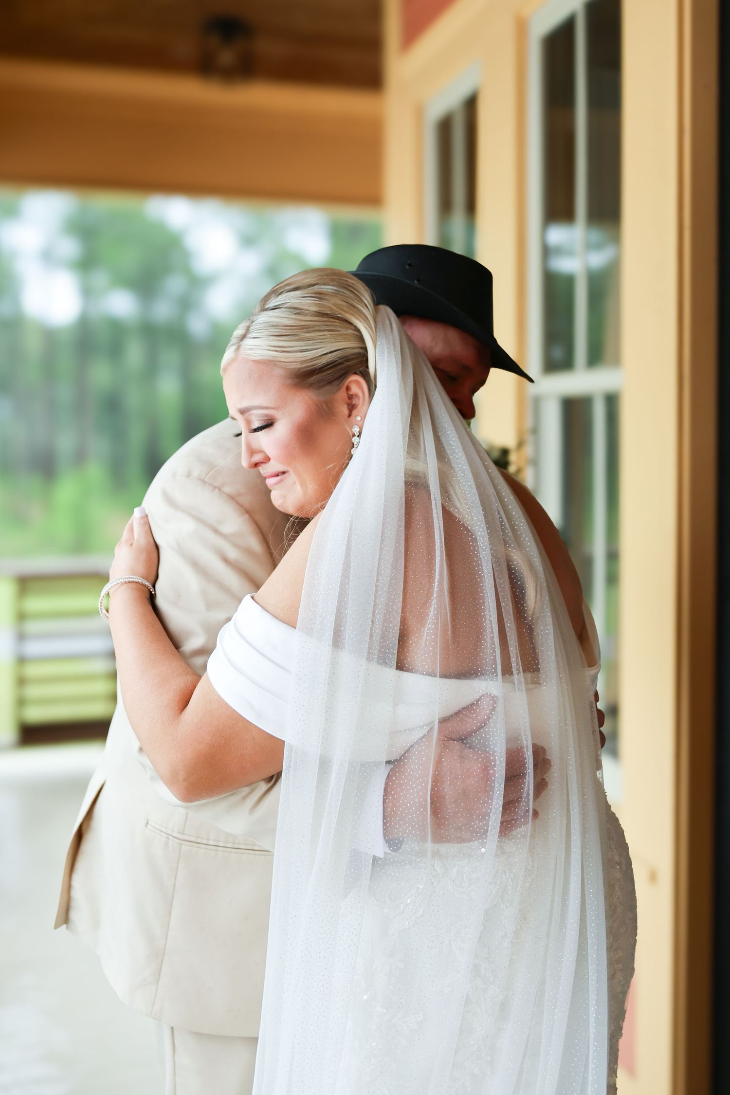Emotional bride in white veil sharing tender hug with father in tan suit and black cowboy hat.